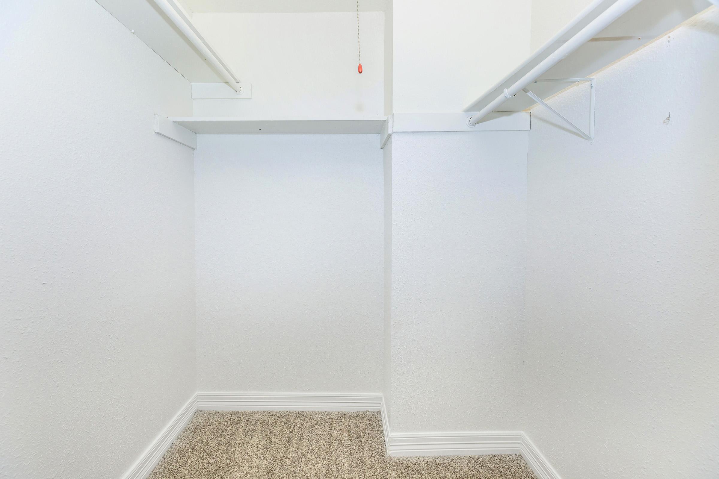 Empty walk-in closet with white walls, a carpeted floor, and two horizontal shelves on either side. A ceiling light fixture hangs above, and there's a pull cord visible. The space appears clean and well-lit, providing ample storage potential.