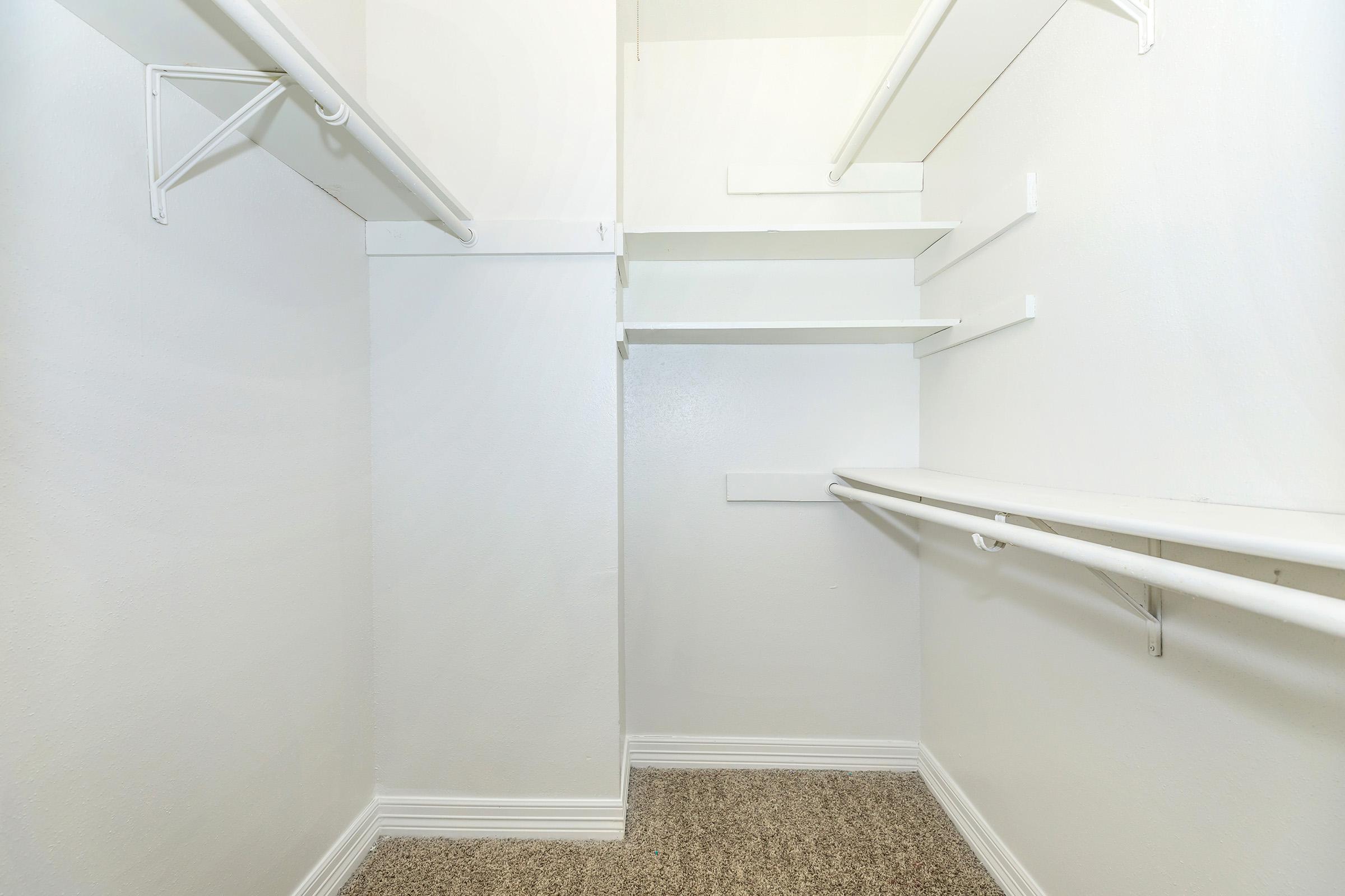 Empty walk-in closet featuring white walls, a light carpeted floor, and multiple shelves and hanging rods for storage. The space appears clean and organized, with a minimalistic design.