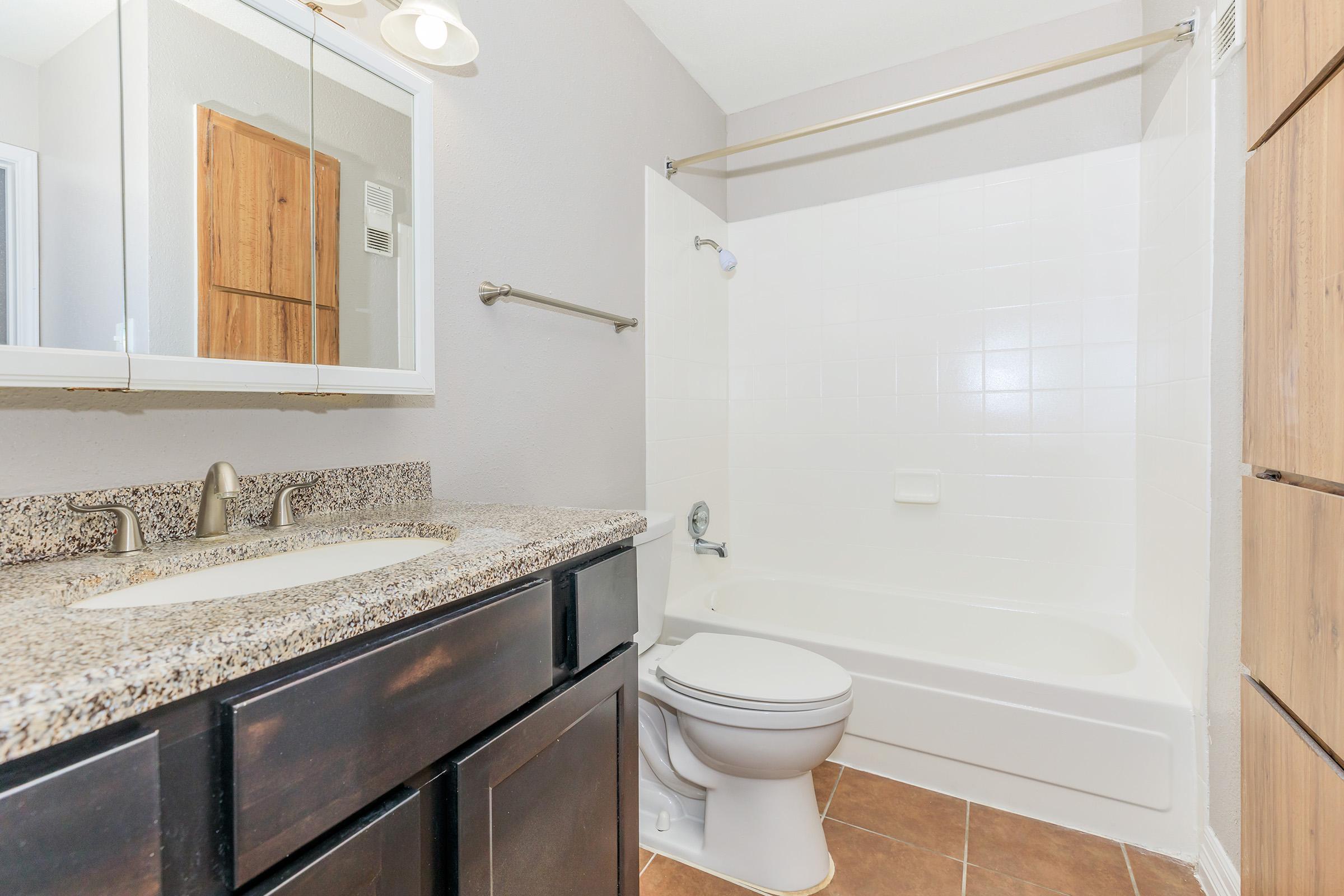 A clean and modern bathroom featuring a white bathtub and shower, a dark wood vanity with a granite countertop, a mirror above the sink, and neutral-colored walls. The tile floor adds a contemporary touch to the space.