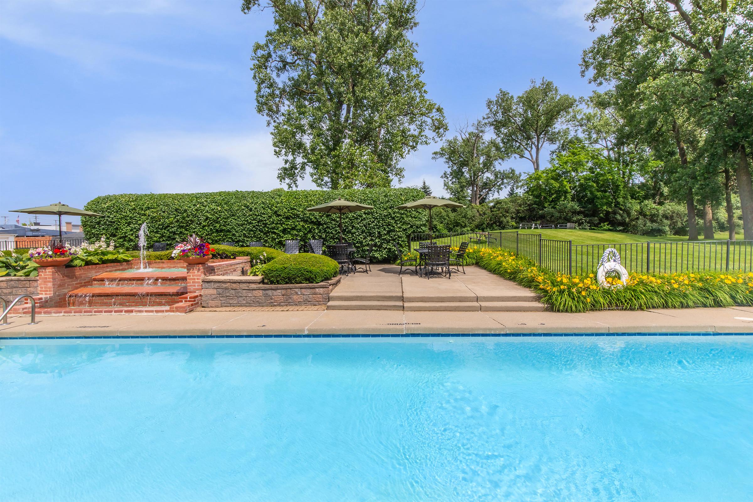 Outdoor pool area featuring a clear blue swimming pool with surrounded landscaping. There are comfortable lounge chairs under umbrellas, a stone wall with decorative planters, and a lush green hedge in the background, creating a serene and inviting atmosphere for relaxation.