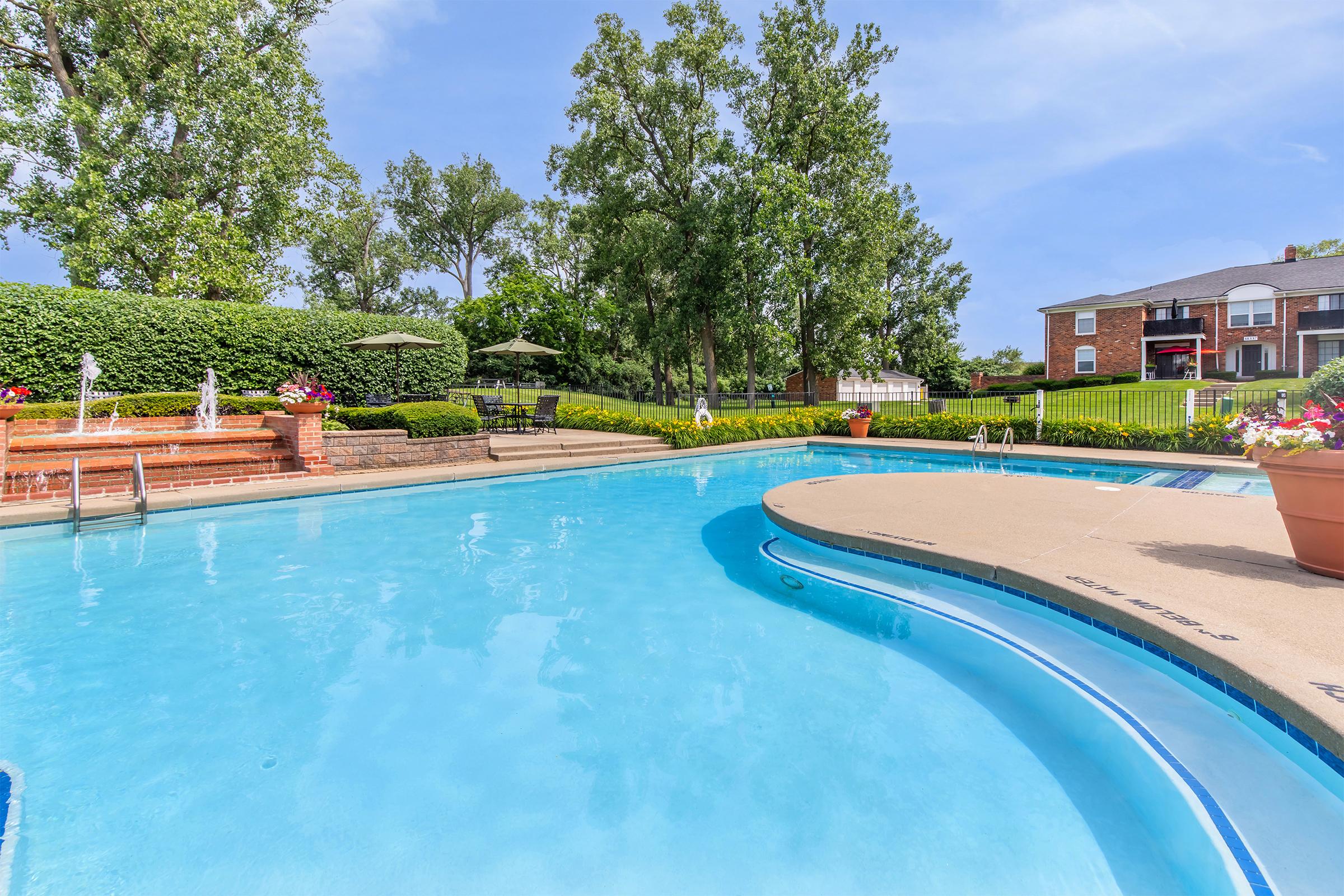 A clear, blue swimming pool surrounded by green trees and well-maintained landscaping. Lounge chairs and umbrellas are positioned around the pool area. In the background, a brick building can be seen with a manicured lawn, creating a relaxing outdoor atmosphere.