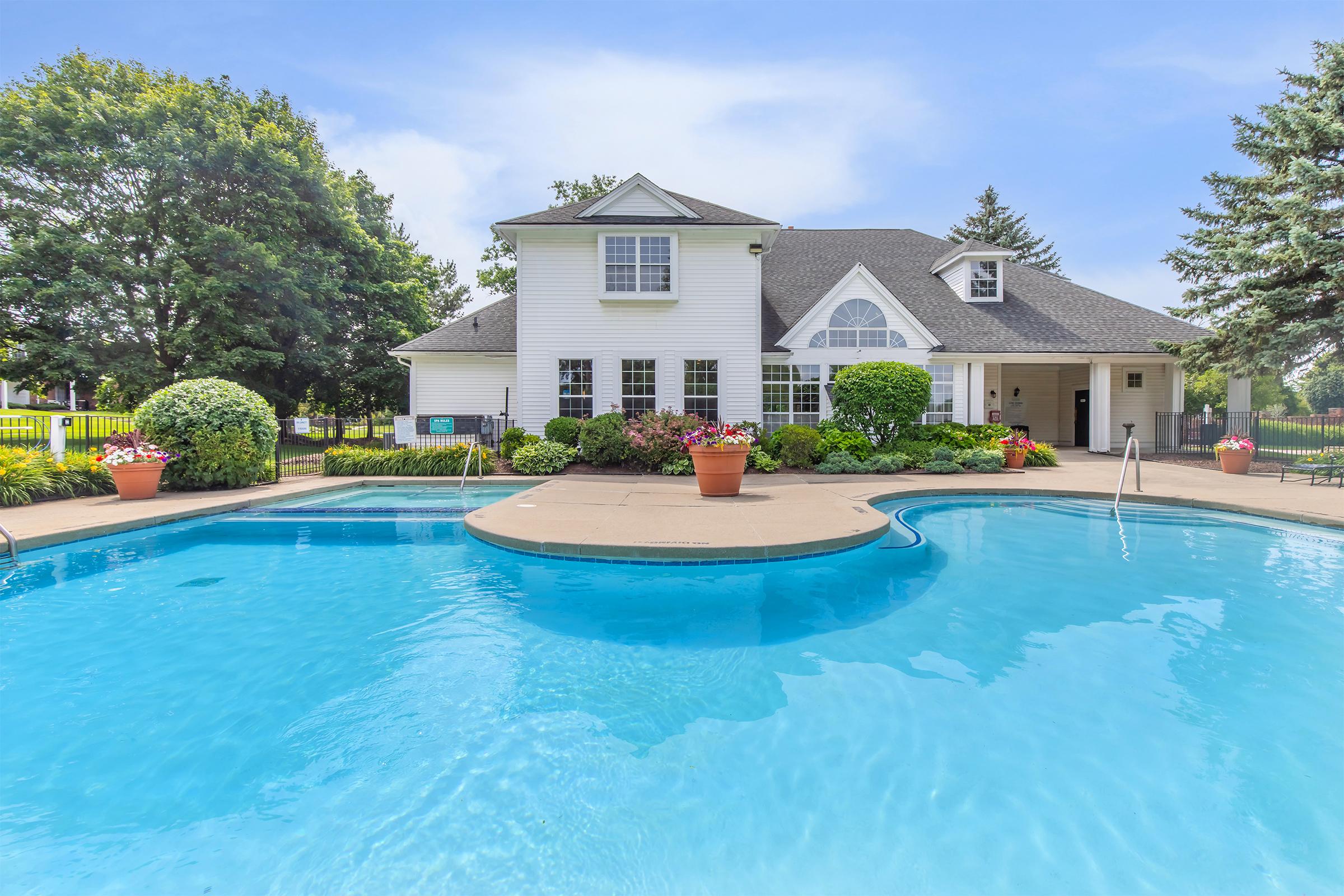 A beautiful residential backyard featuring a large, sparkling blue swimming pool with a circular deck. Lush greenery surrounds the pool, with colorful flower pots and landscaping. In the background, a spacious white house with a gabled roof and large windows is visible, set against a clear blue sky.