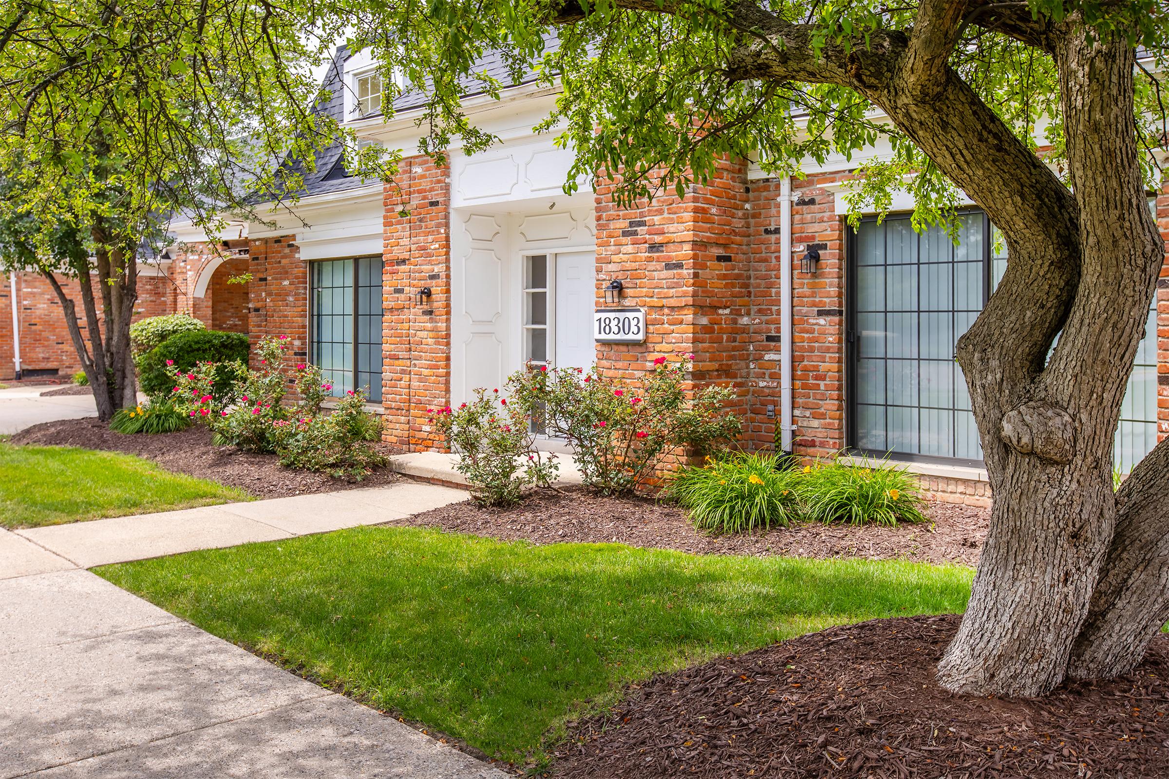 A well-maintained brick building with a white door and large windows, surrounded by green shrubs and flowering rose bushes. A tree with a thick trunk and lush green leaves is visible in the foreground, alongside a concrete pathway leading up to the entrance. The setting is tidy and inviting.
