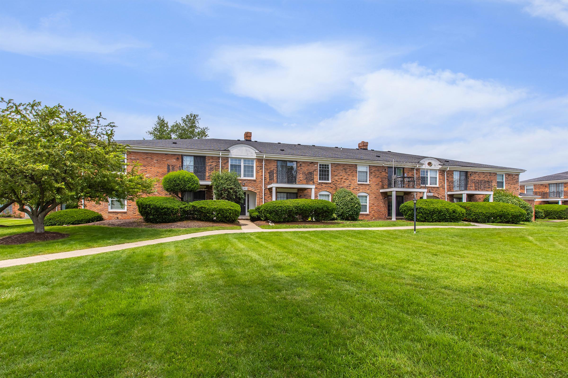 A brick apartment building with multiple units, surrounded by well-maintained green lawns and shrubs. The sky is clear with a few clouds, creating a bright and inviting atmosphere.