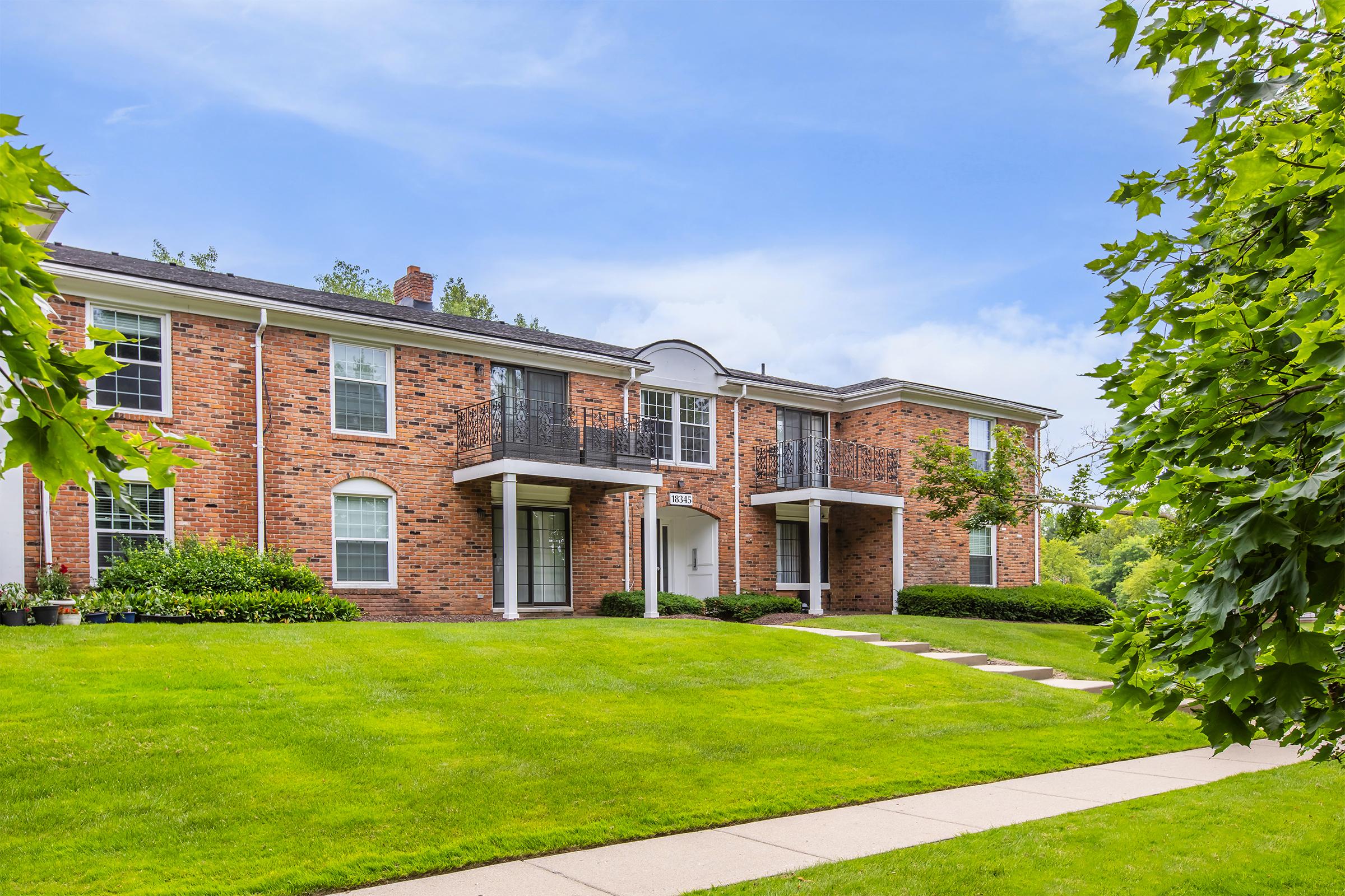 A well-maintained brick apartment building with green lawns and trees surrounding it. It features two entrances with white columns and balconies, set against a clear blue sky. A walkway leads up to the entrances, enhancing the inviting appearance of the property.