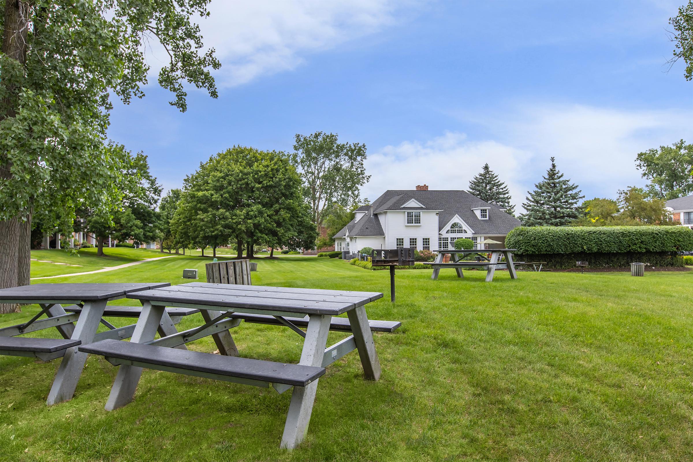A serene park scene featuring several wooden picnic tables in an open green space. Lush grass and trees create a tranquil atmosphere, with a white house and various shrubs in the background under a partly cloudy sky. The setting is ideal for outdoor gatherings or relaxation.