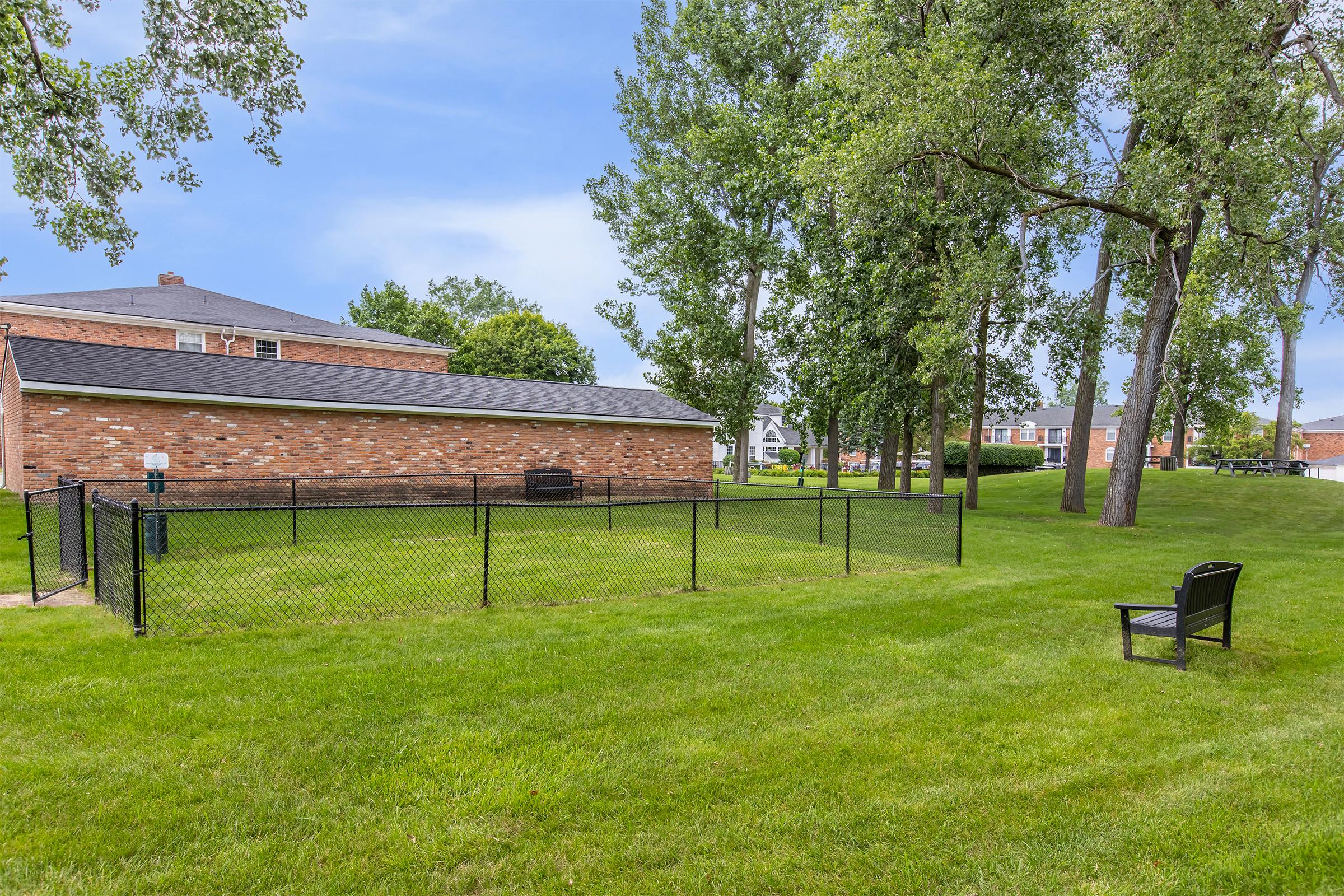 A grassy area enclosed by a black chain-link fence, featuring a small wooden chair on the lawn. In the background, a brick building is partially visible along with a row of trees and neighboring structures. The sky is clear, suggesting a sunny day.