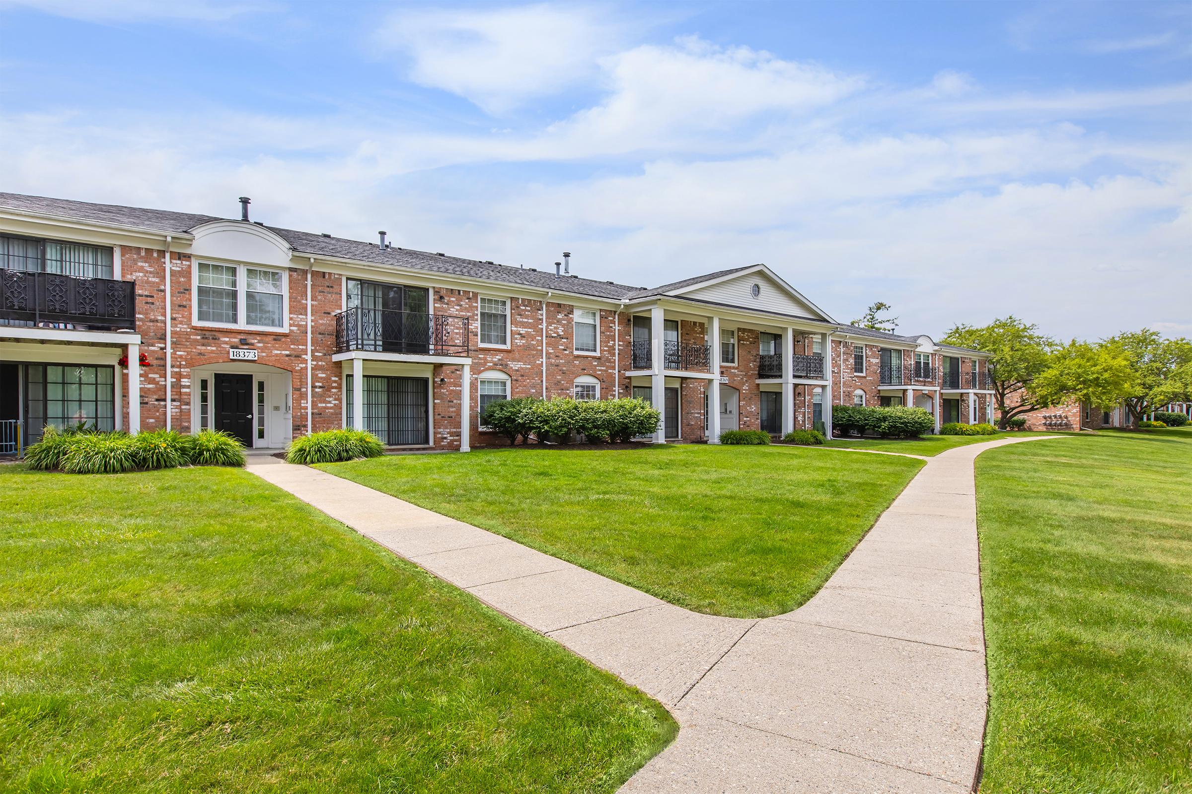 Row of brick apartment buildings with balconies, situated along a grassy path in a well-maintained landscape. Lush green lawns surround the buildings under a partly cloudy sky.