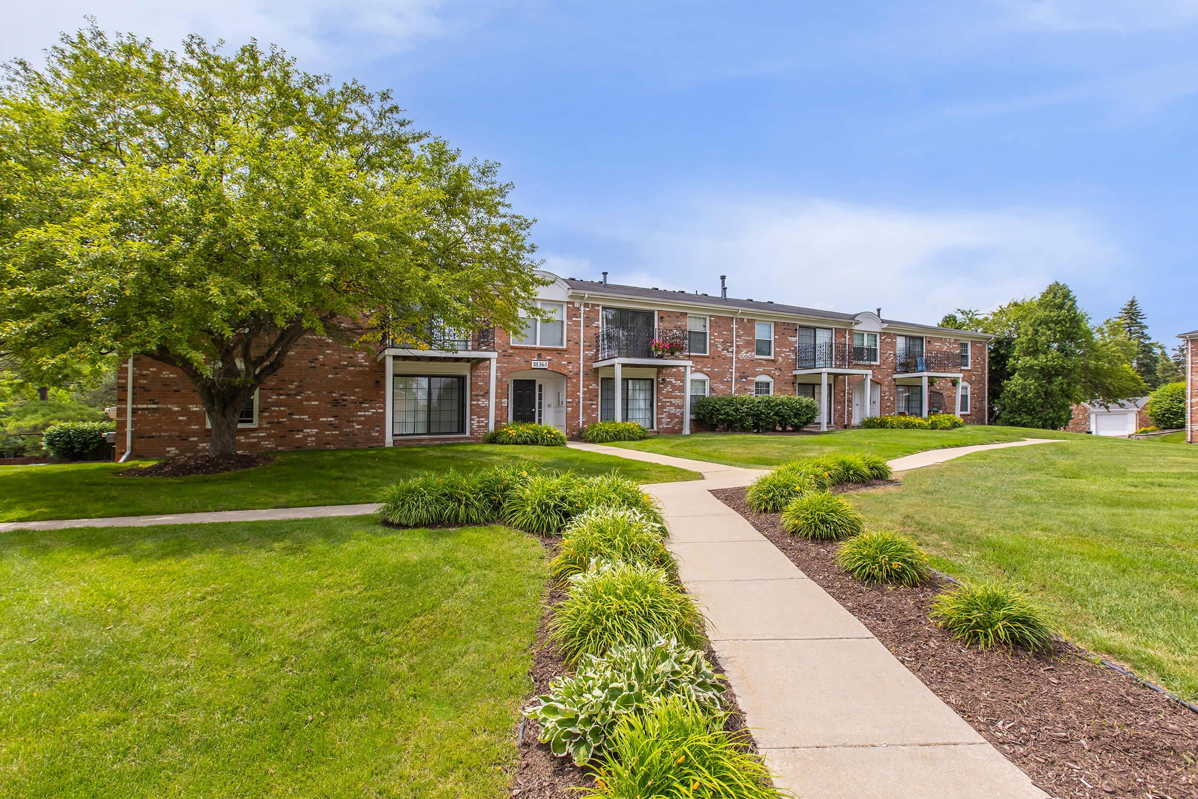 A well-maintained apartment complex featuring brick buildings surrounded by lush green lawns and well-trimmed shrubs. Pathways meander through the landscape, leading to the entrances of the apartments. Trees provide shade, enhancing the inviting atmosphere of the property.