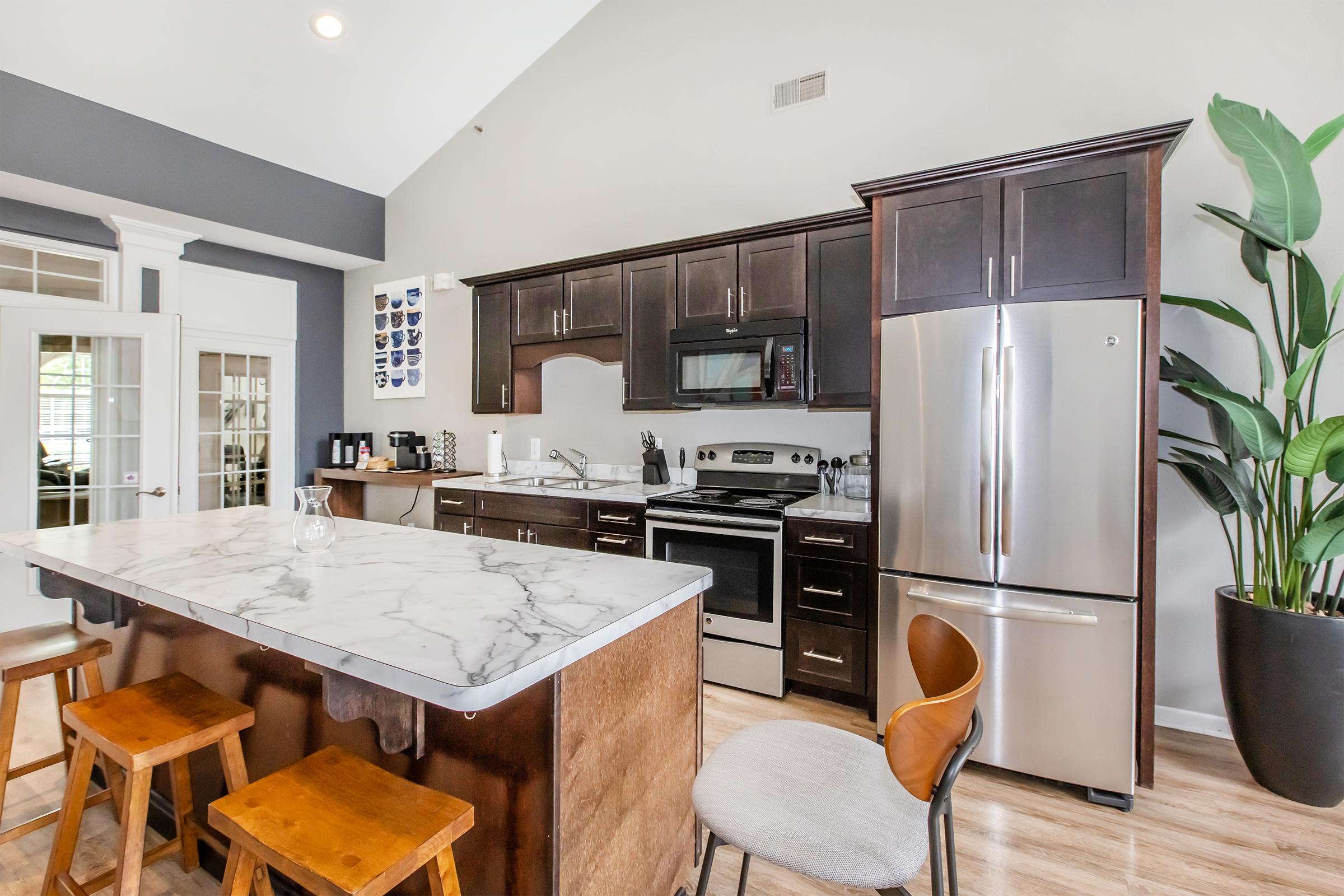 A modern kitchen featuring dark wood cabinets, stainless steel appliances, and a marble-topped island with wooden stools. The space is well-lit with natural light and has a high ceiling, large windows, and a houseplant in the corner.
