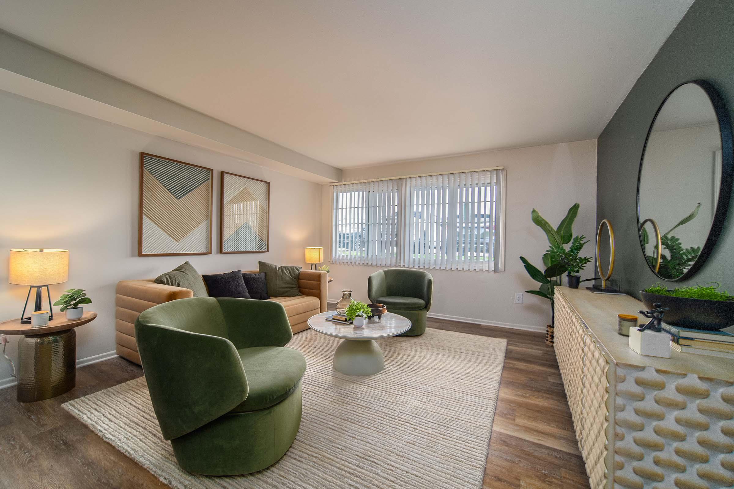 A modern living room featuring two green upholstered chairs, a round coffee table, and a textured area rug. There are decorative plants and a side table with a lamp, along with two geometric art pieces on the wall and a large round mirror above a console table. Bright natural light enters through window blinds.