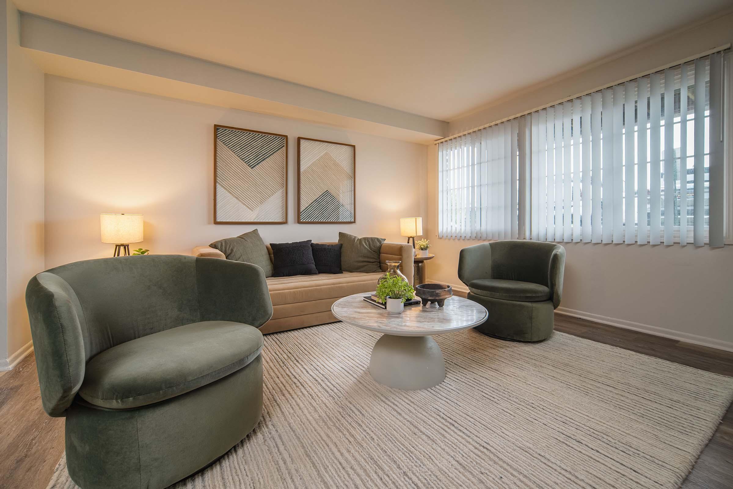 A cozy living room featuring two round green chairs and a circular marble coffee table on a light rug. The space includes a beige sofa against a wall, two modern art pieces above the sofa, and a lamp on a side table. Large windows with white blinds allow natural light to illuminate the room.