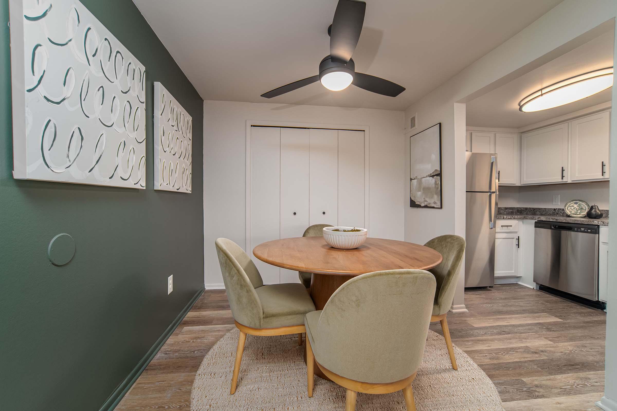 A cozy dining area featuring a round wooden table surrounded by four plush green chairs. The walls are painted a dark green with abstract white artwork, and there is a ceiling fan above. A closet with white sliding doors is visible, along with a kitchen area in the background that includes stainless steel appliances.