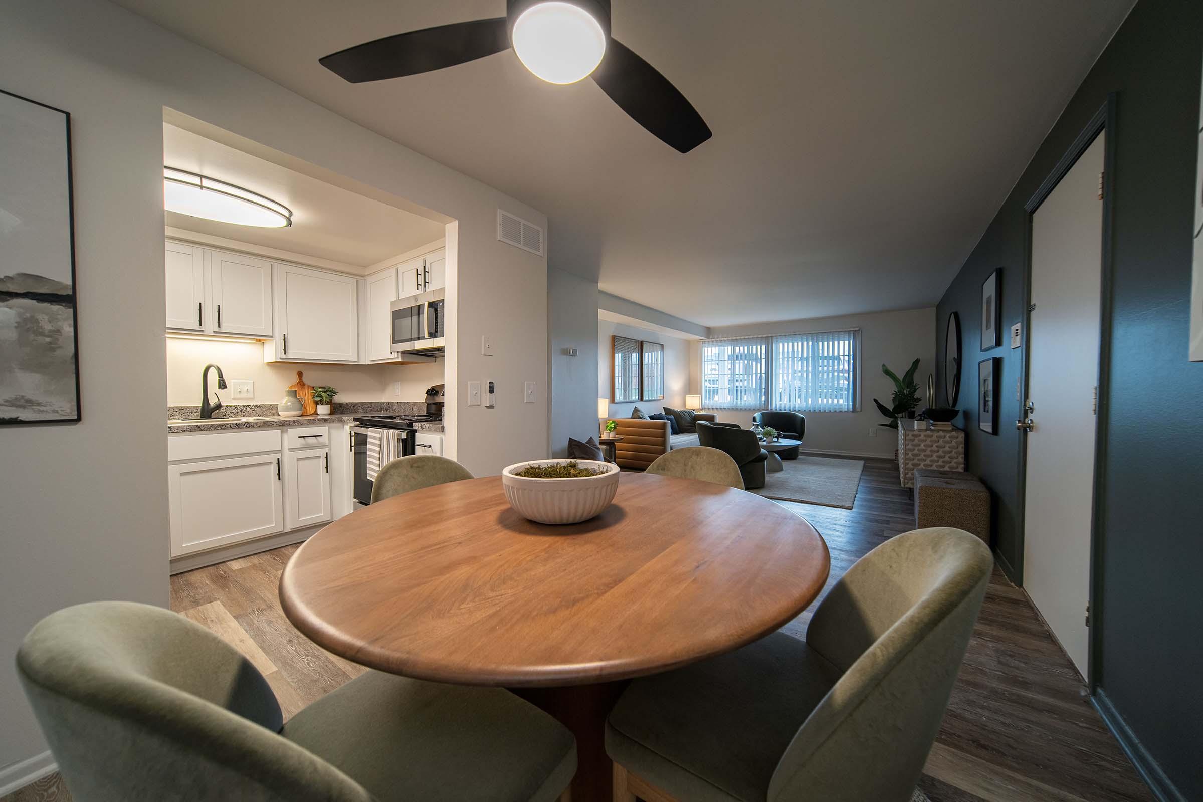 A modern dining area featuring a round wooden table with green chairs. In the background, a stylish kitchen is visible, along with a cozy living room space that includes a sofa and large windows. The decor is contemporary, with warm colors and well-arranged furnishings.