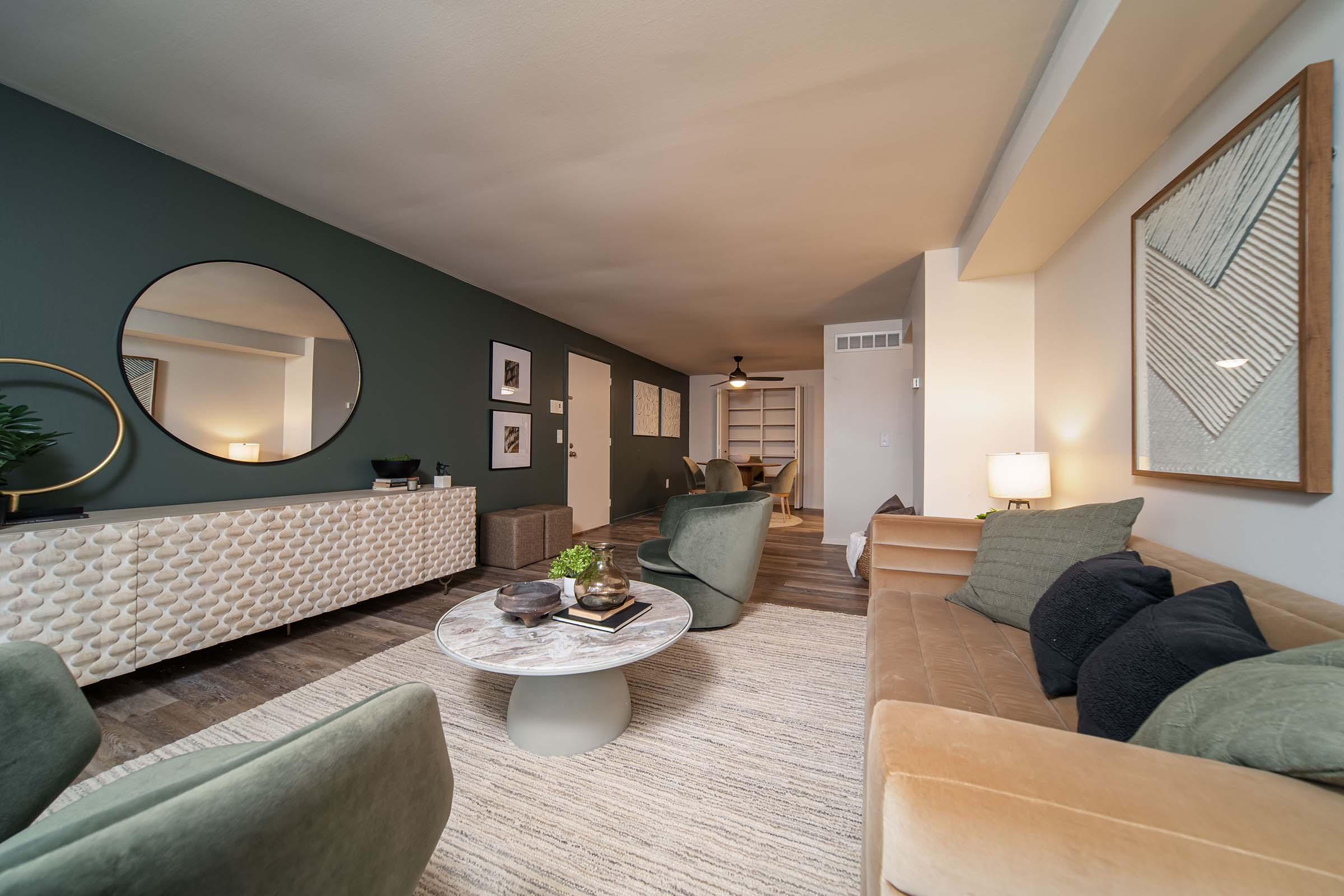 A modern living room featuring a stylish design with a round mirror on the wall, a light-colored sofa, green accent chairs, and a circular coffee table. The decor includes framed artwork, a sideboard, and a neutral-toned area rug, creating a cozy and inviting atmosphere.