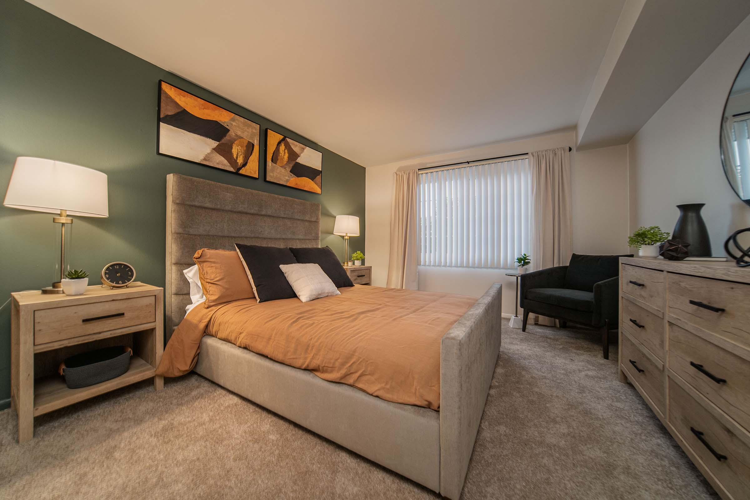 A cozy bedroom featuring a plush bed with brown and white bedding, two bedside tables with lamps, abstract artwork on the wall, a large window with sheer curtains, and a small black armchair. The walls are painted a muted green, and there are decorative plants on the furniture.