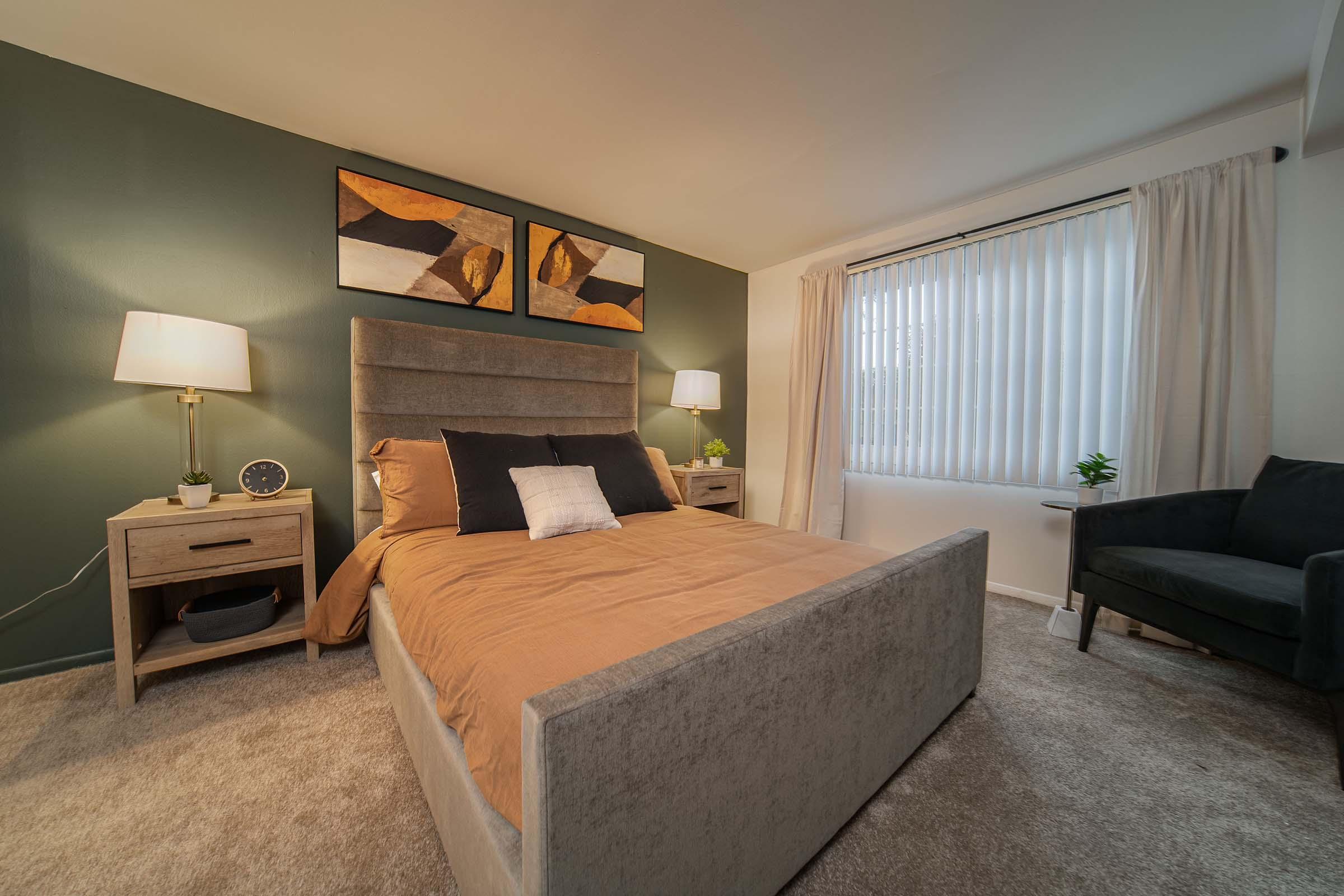 A cozy bedroom featuring a stylish bed with a beige comforter and decorative pillows. On either side of the bed, there are modern nightstands with lamps. Above the bed, two abstract art pieces hang on the wall. A large window with blinds allows natural light to enter, and a small plant adds a touch of greenery.