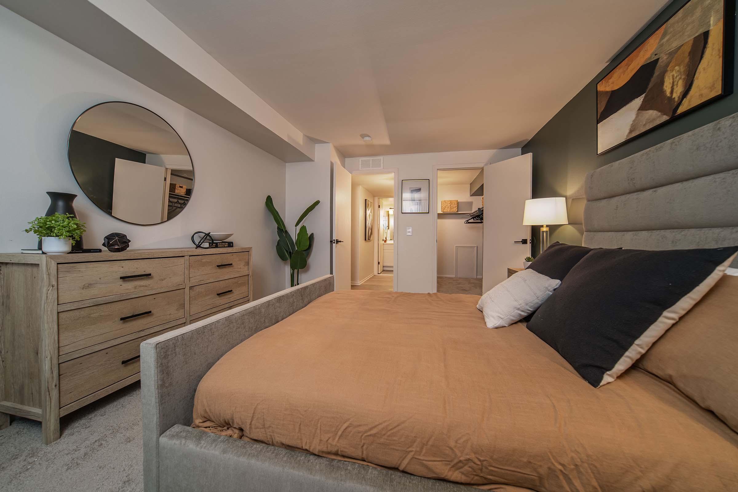 A cozy bedroom featuring a plush bed with an orange-brown duvet and decorative pillows. A wooden dresser sits against one wall, and a round mirror hangs above it. A plant adds a touch of greenery. A modern framed artwork decorates the wall, and a closet is visible in the background, contributing to a warm and inviting atmosphere.