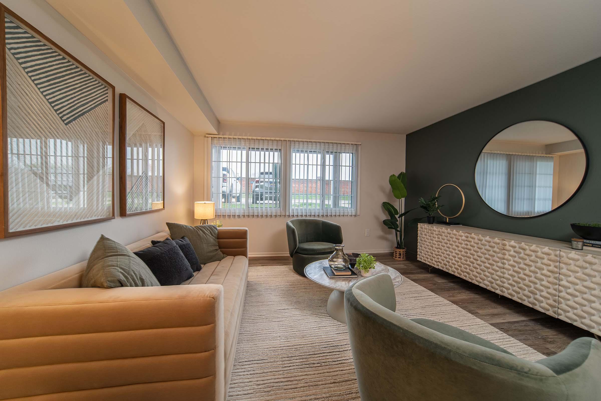 A modern living room featuring a beige sofa with throw pillows, two green lounge chairs, a round mirror on the wall, and decorative framed artwork. Natural light streams in through large windows, illuminating a stylish sideboard and a small potted plant. The flooring is a mix of textures with a light area rug.