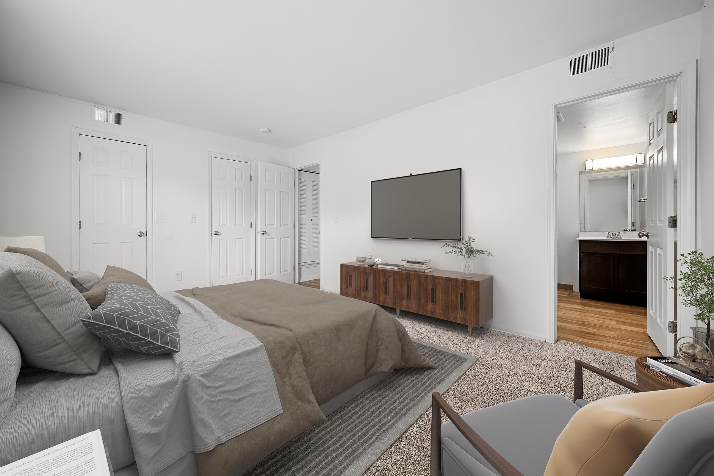 A spacious and modern bedroom featuring a large bed with neutral-colored bedding, a wood media console with a TV mounted on the wall, and a cozy armchair. A bathroom is visible through an open door, and the room has light-colored walls and a plush carpet. Natural light brightens the space.