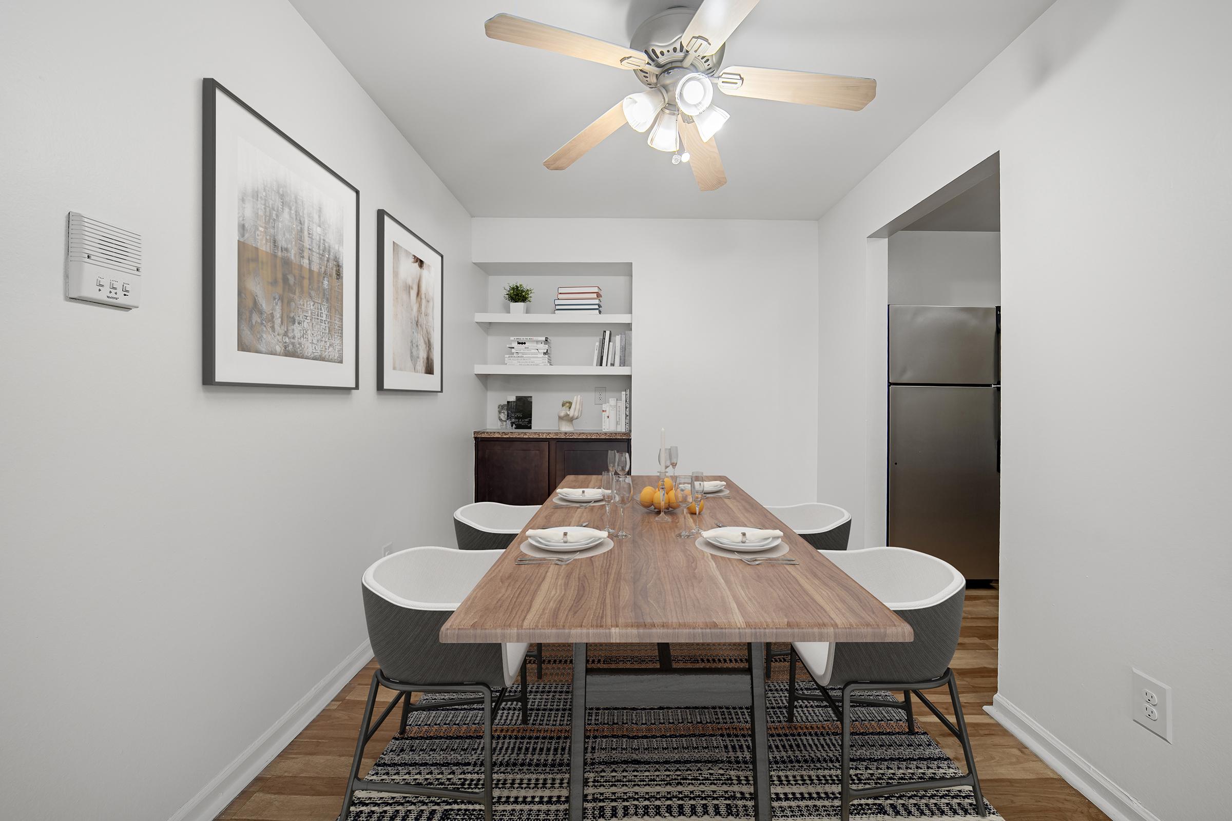 A modern dining area features a wooden table set for four with plates and glasses. A small bowl of oranges sits in the center. The walls are adorned with framed artwork, and a shelf with decor items is visible in the background. A ceiling fan adds a functional touch to the bright, neutral-colored space.