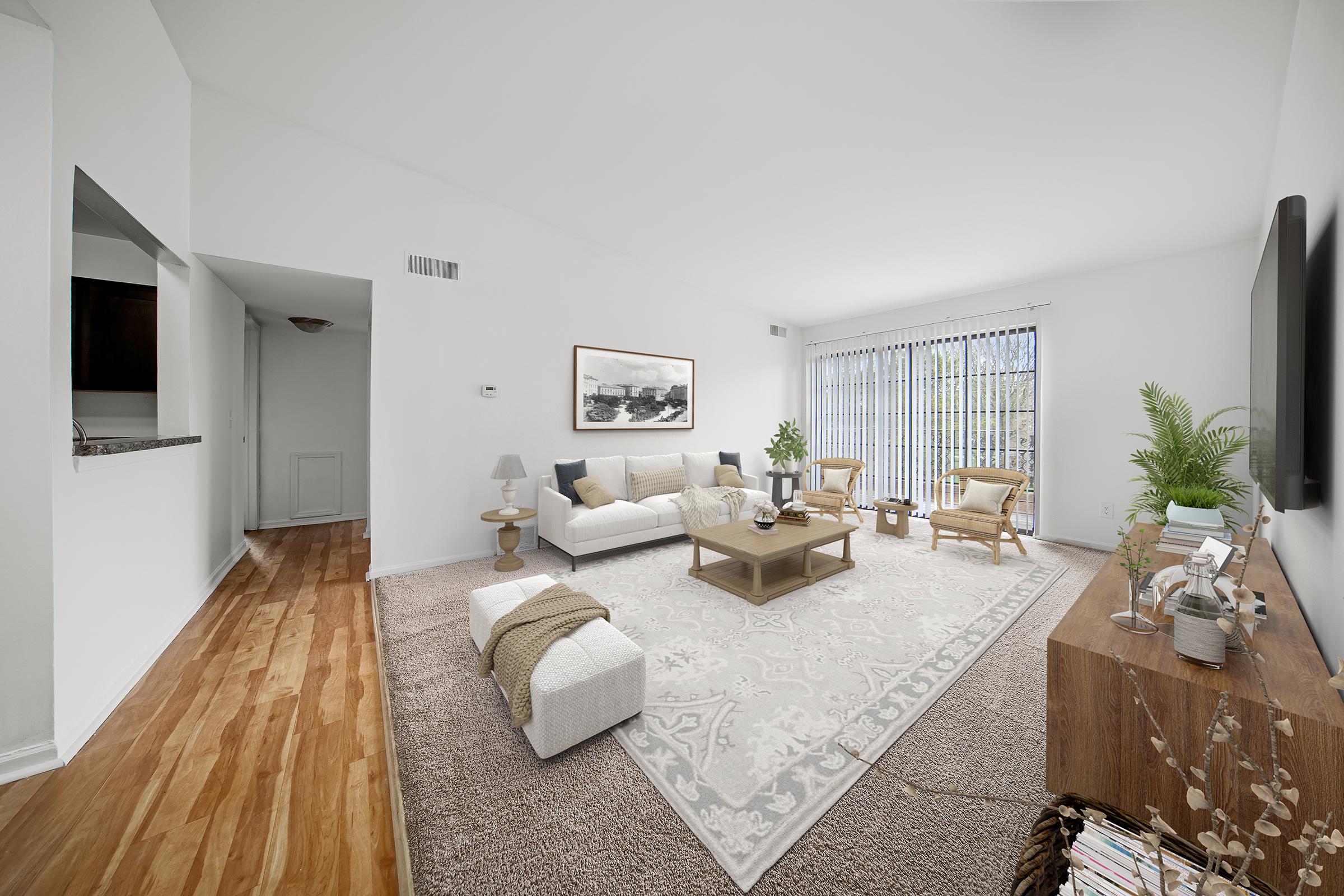 A bright, spacious living room with a light color scheme. It features a white sofa with decorative cushions, a wooden coffee table, and two stylish chairs. Large windows allow natural light to flood the space, with greenery visible outside. A cozy rug and a modern TV unit complete the inviting atmosphere.