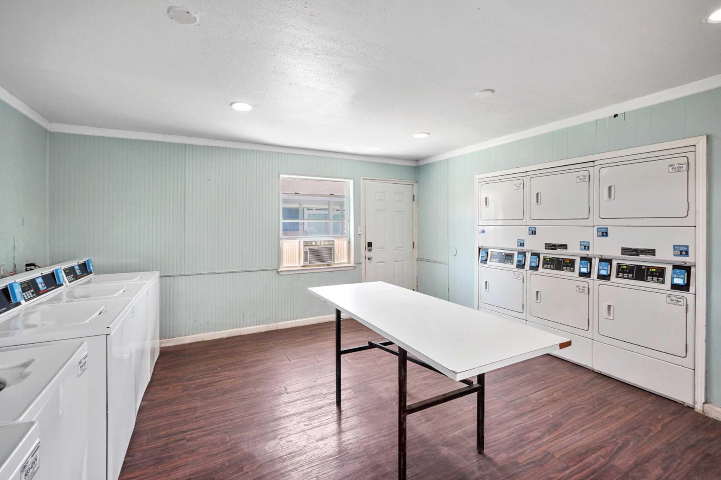 A clean, well-lit laundry room featuring several white washing machines and dryers lined against the wall. In the center, there’s a white folding table. A window allows natural light to enter, and there’s an air conditioning unit mounted near the window. The walls are painted light green, and the floor is dark wood.