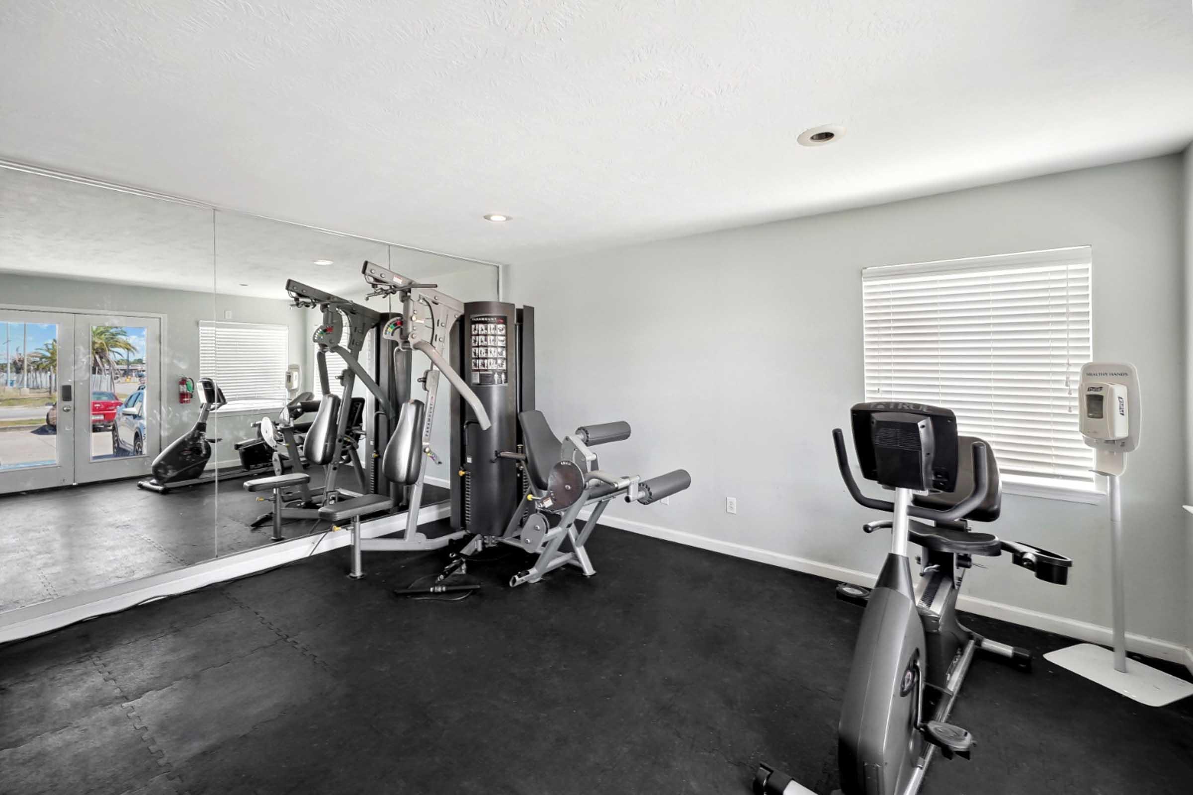 A modern gym interior featuring various exercise equipment, including a multi-station weight machine, an upright bike, and space for workouts. The room has large mirrors reflecting the equipment, and a window allowing natural light to enter, creating a bright and inviting atmosphere for fitness activities.