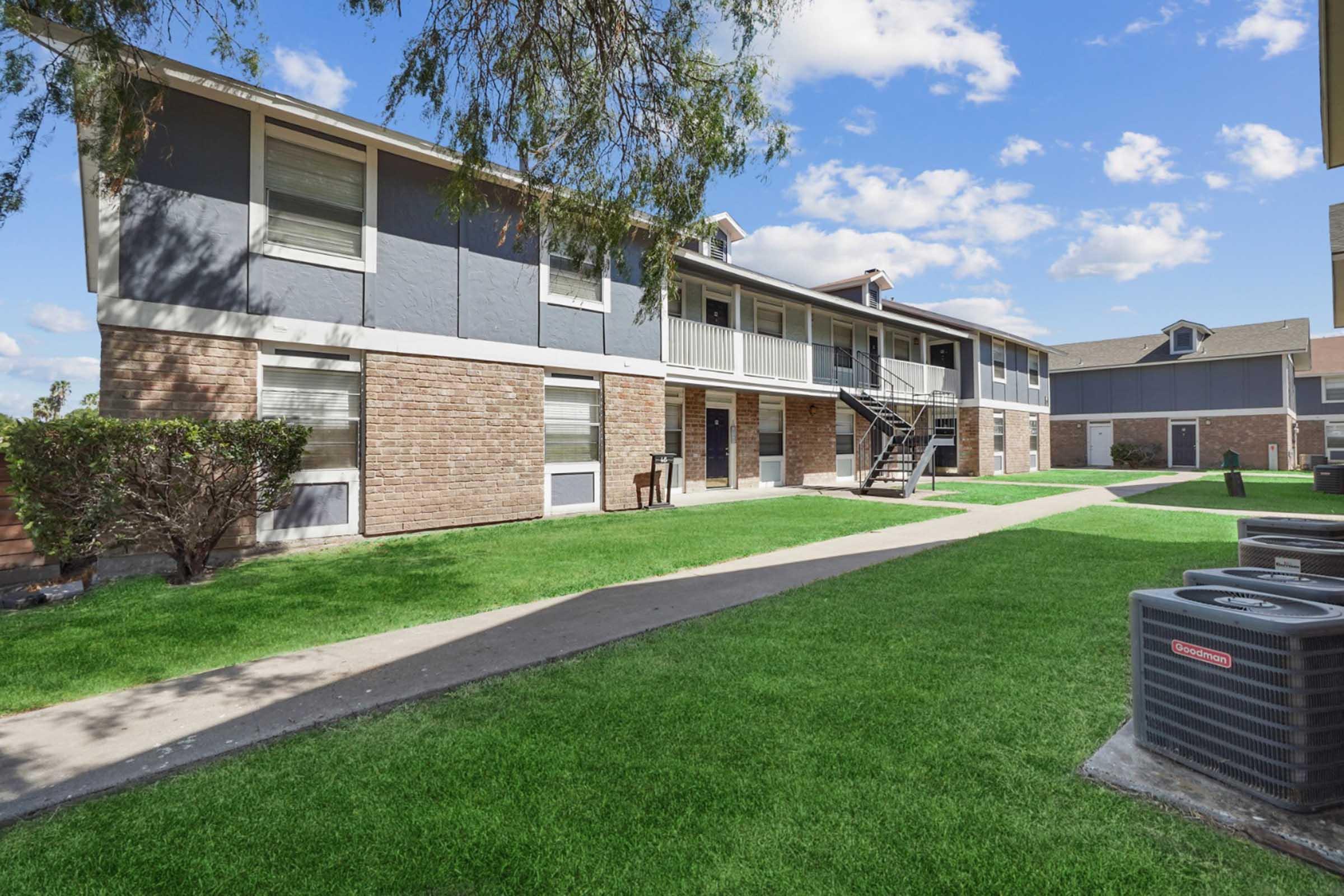 Two-story residential building with gray and brick exterior, featuring multiple balconies. A well-maintained green lawn surrounds the building, with a sidewalk running through the grass. In the foreground, there is an outdoor air conditioning unit. The sky is partly cloudy.