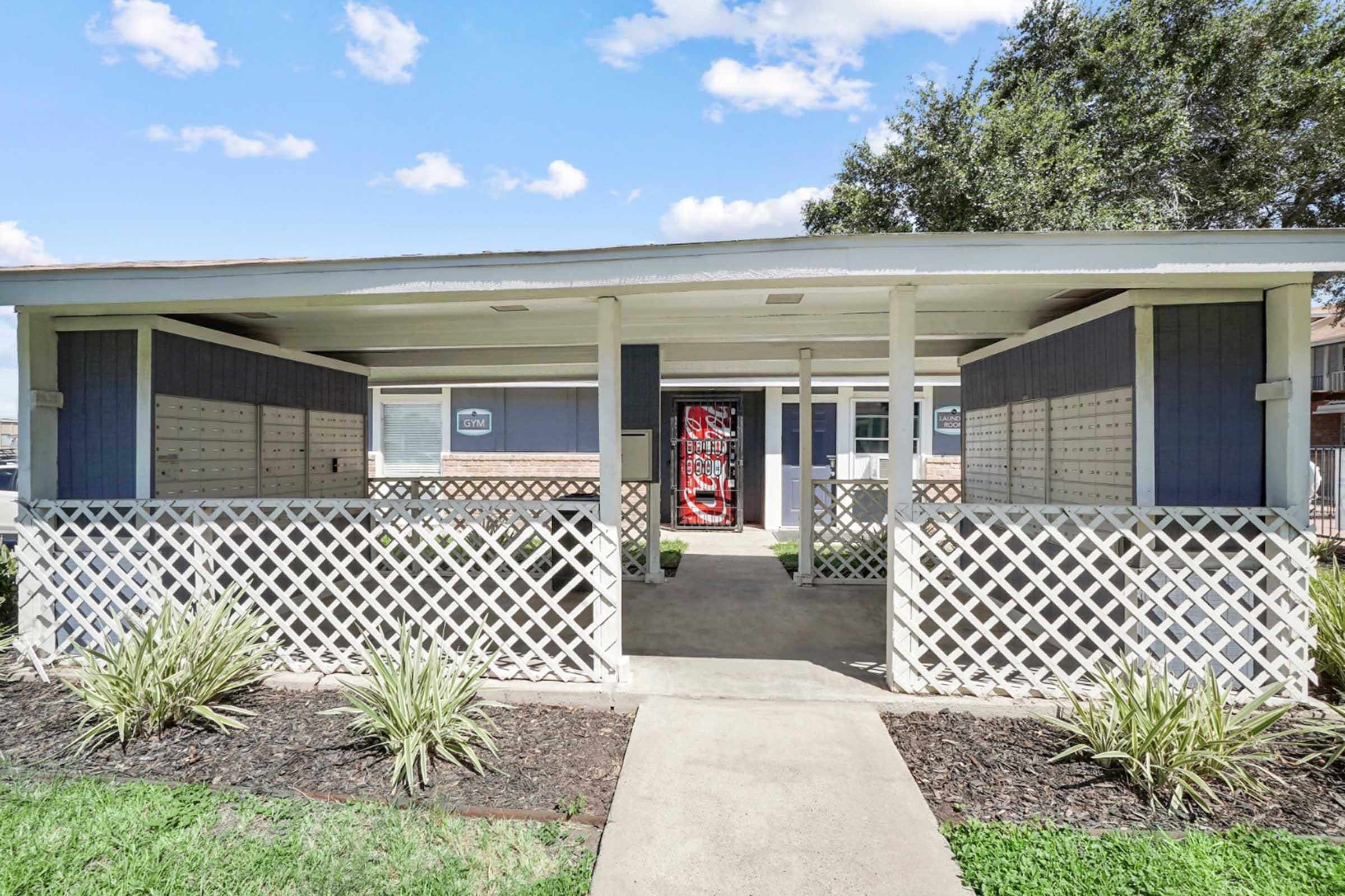 A small building featuring a covered porch with decorative lattice work, mailboxes on the walls, and a colorful door at the center. Surrounding the entrance are landscaped areas with low plants and freshly cut grass under a blue sky with fluffy clouds.