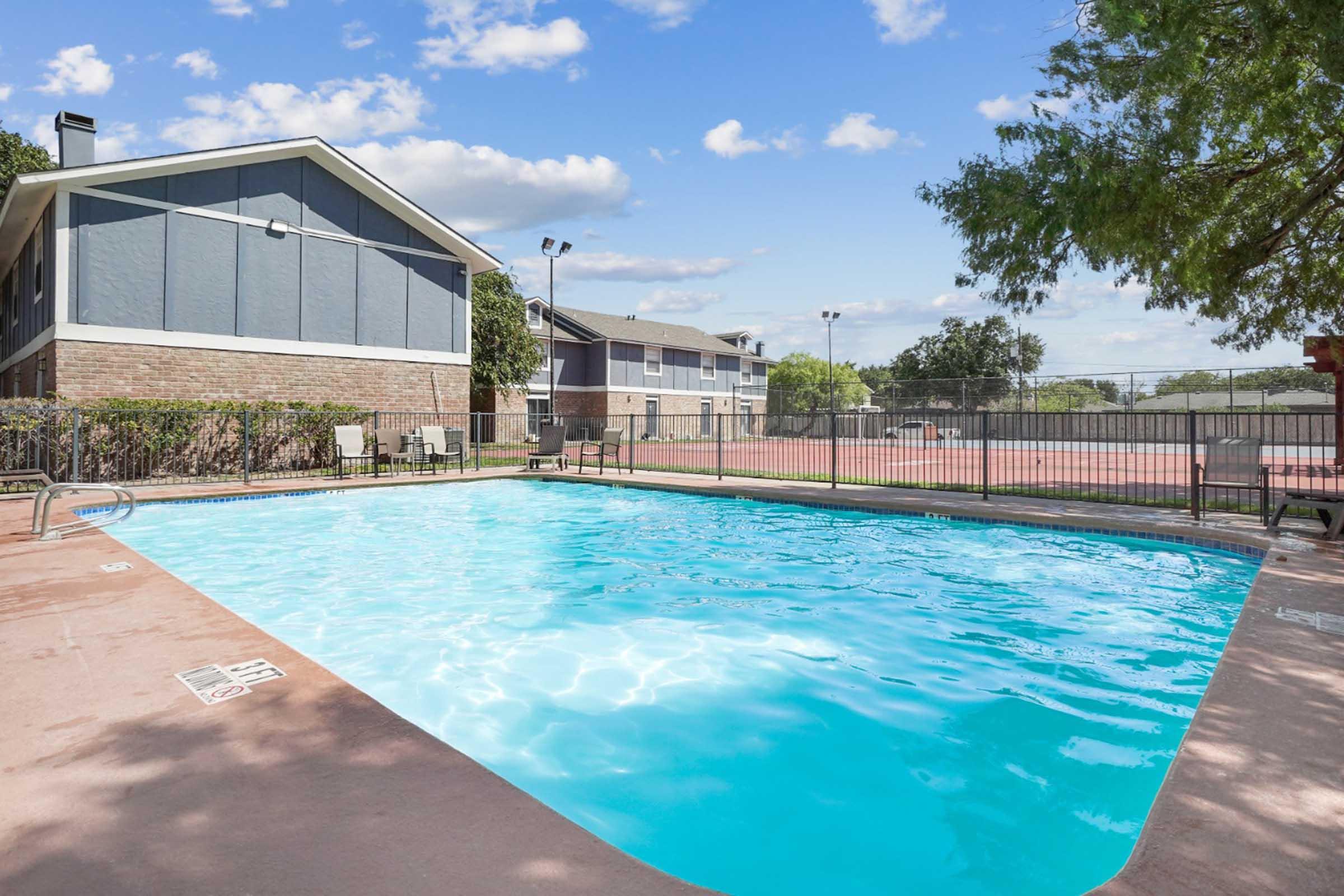 A swimming pool surrounded by a fence, with clear blue water. In the background, there are several residential buildings and a tennis court. The sky is bright with a few clouds, and greenery lines the pool area, creating a vibrant, inviting atmosphere.