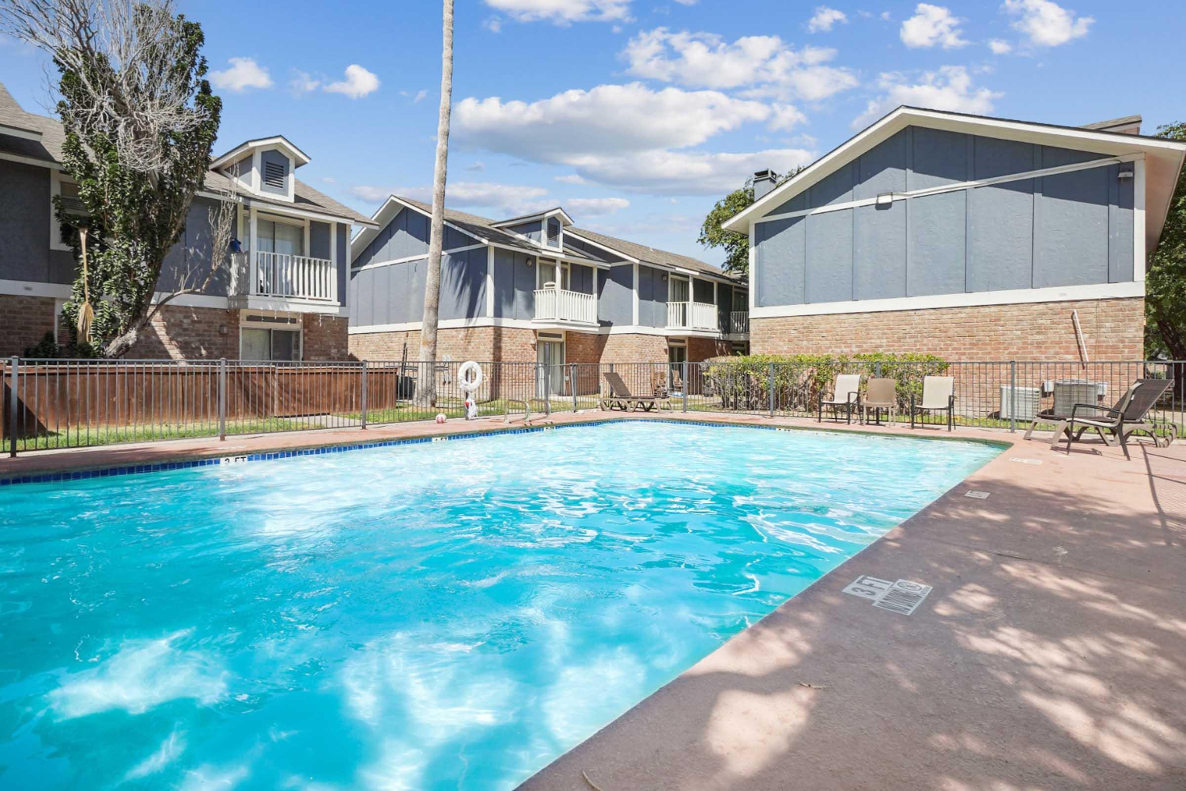 A clear swimming pool surrounded by a fenced area, with lounge chairs nearby. In the background, there are two apartment buildings with a mix of brick and siding exteriors. The sky is partly cloudy, creating a bright and inviting atmosphere.