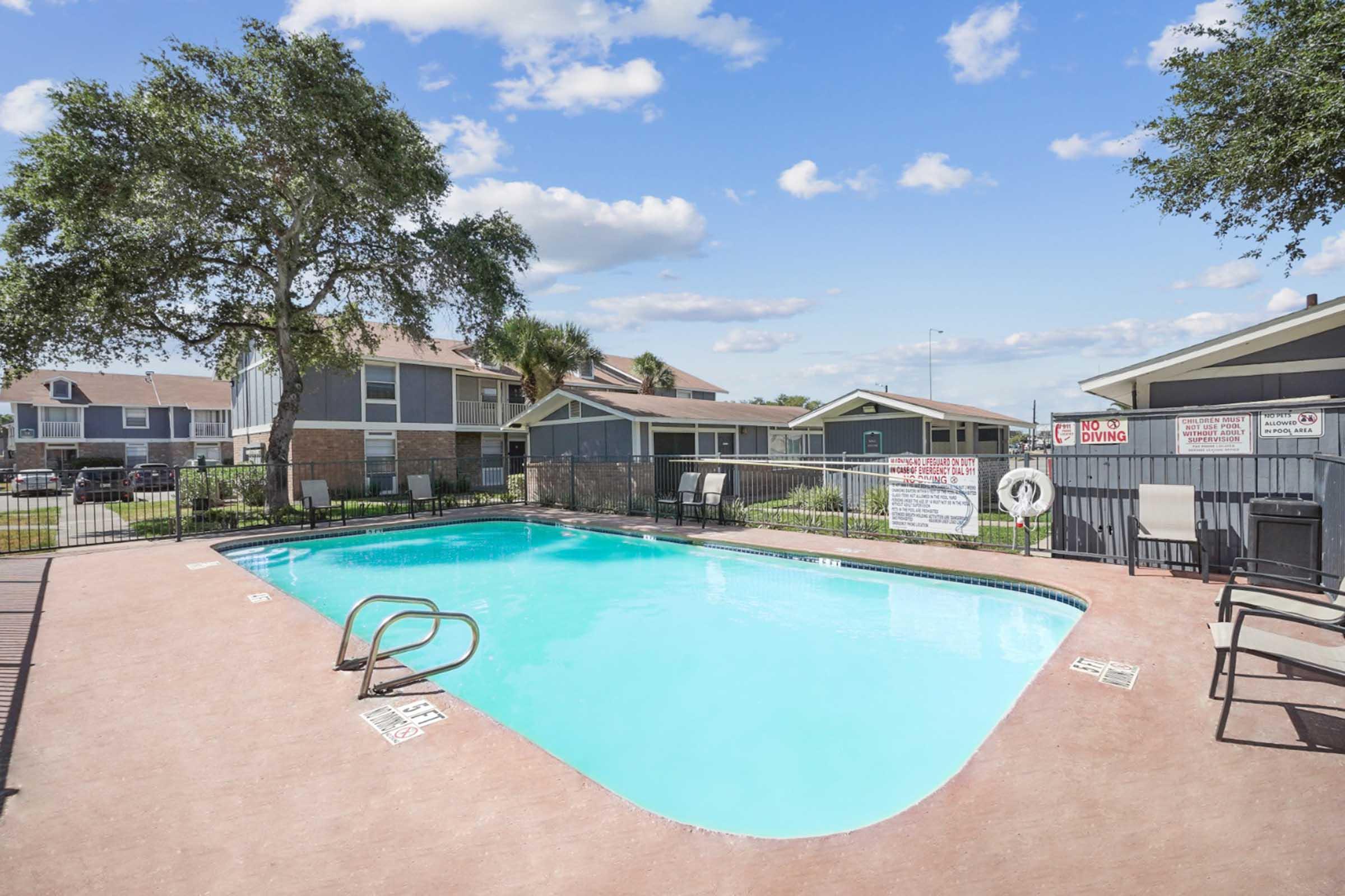A clear swimming pool surrounded by a patio area with lounge chairs. The background features residential buildings and a grassy area. Signs are posted by the pool area, and a tree provides some shade. The sky is blue with scattered clouds.