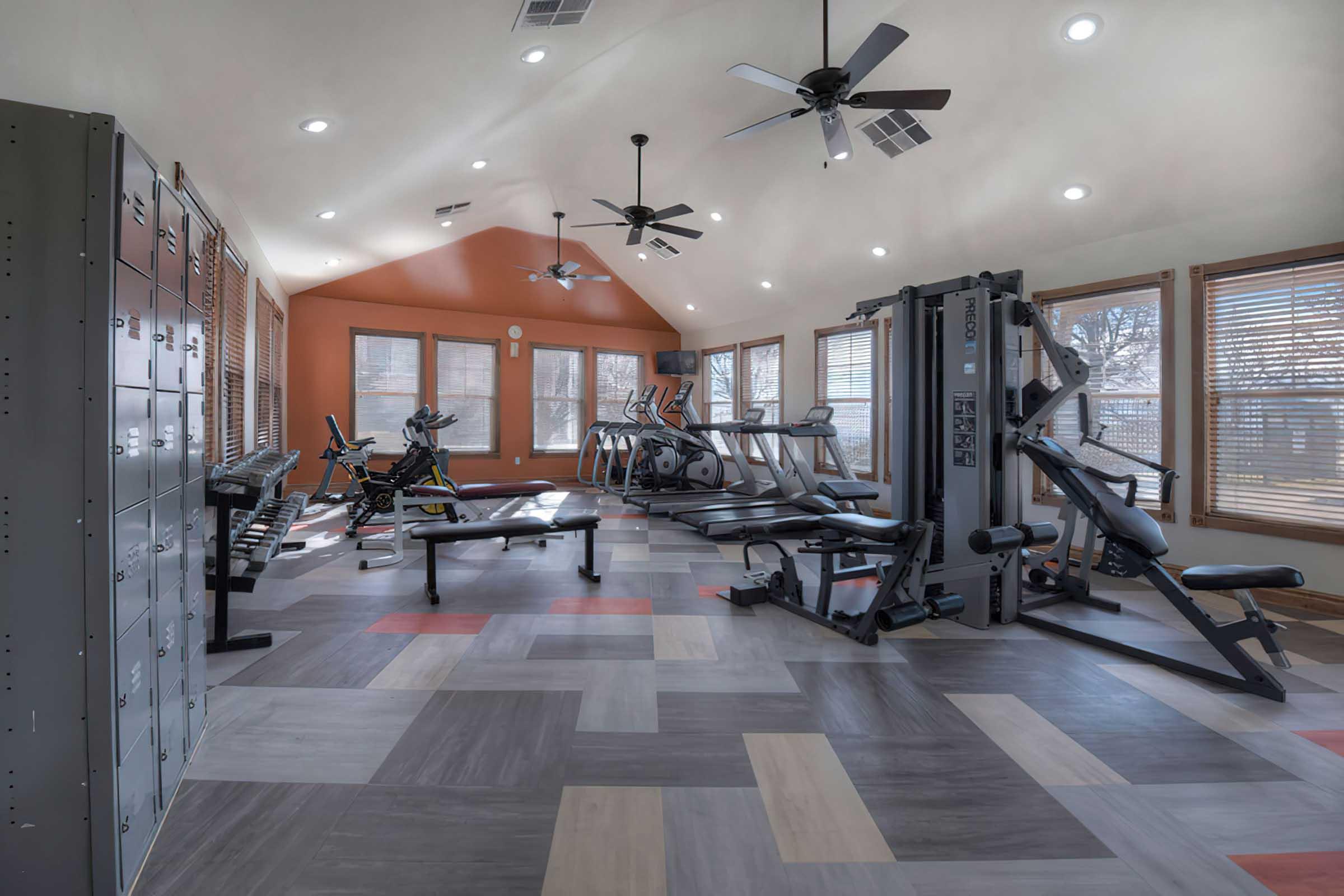 A spacious, well-lit gym featuring multiple exercise machines, weight benches, and lockers. The room has large windows providing natural light and a modern design with colorful flooring. Ceiling fans and bright lights enhance the workout environment.
