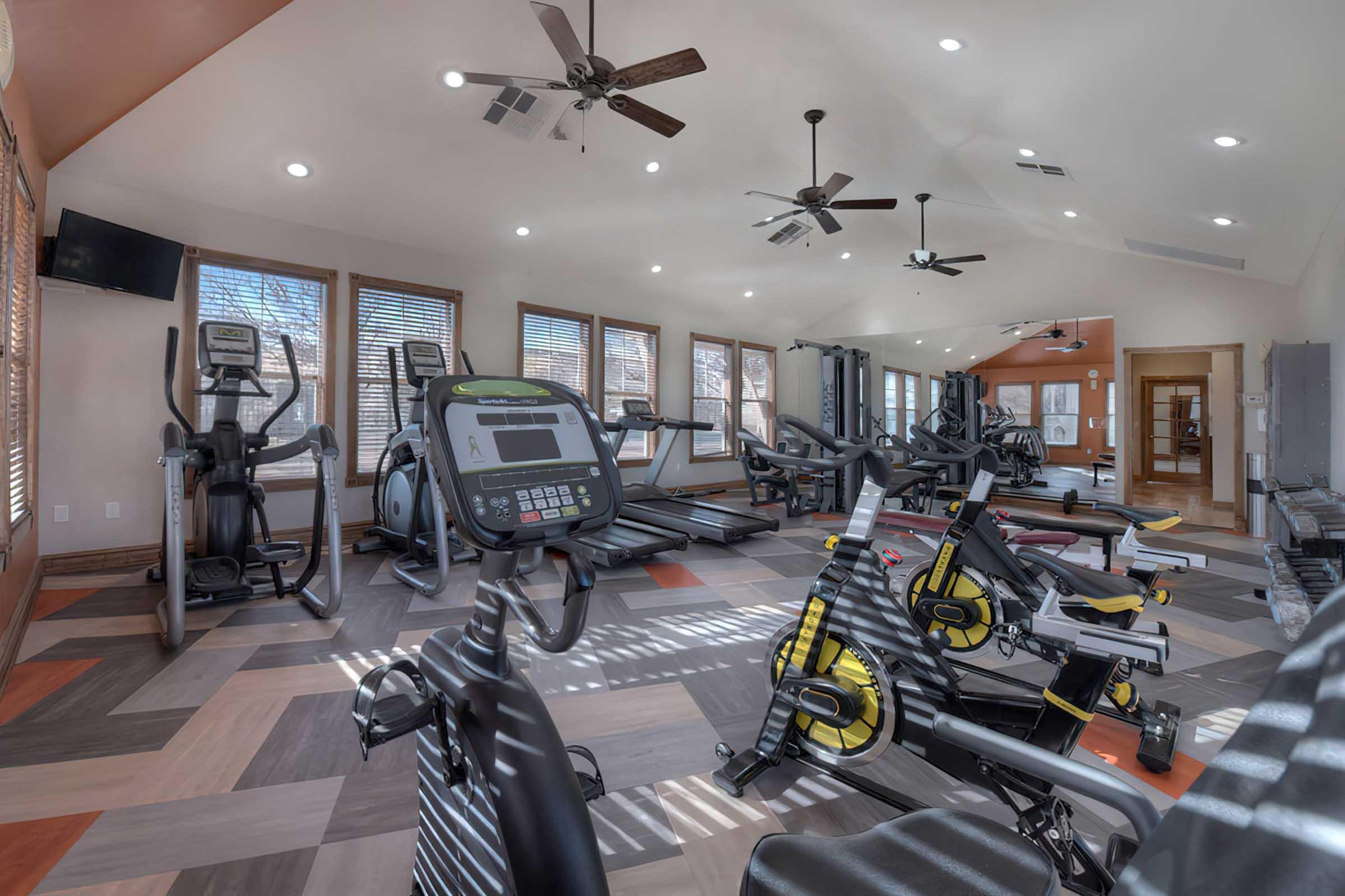 A spacious gym with various exercise equipment, including treadmills, stationary bikes, and elliptical machines. The room features large windows allowing natural light, modern lighting fixtures, and a colorful patterned floor. Several ceiling fans are installed for ventilation.