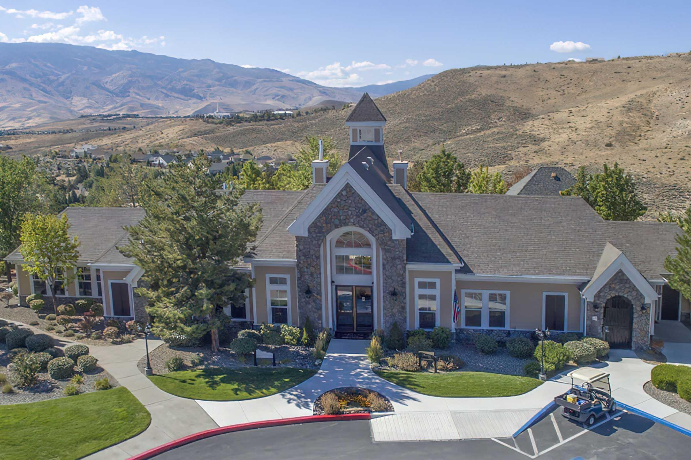 A large building with a stone facade and a tower, surrounded by landscaped gardens and pathways. There are trees and shrubs in the foreground, with a mountainous landscape in the background under a clear blue sky. A golf cart is parked in the foreground along a red-lined driveway