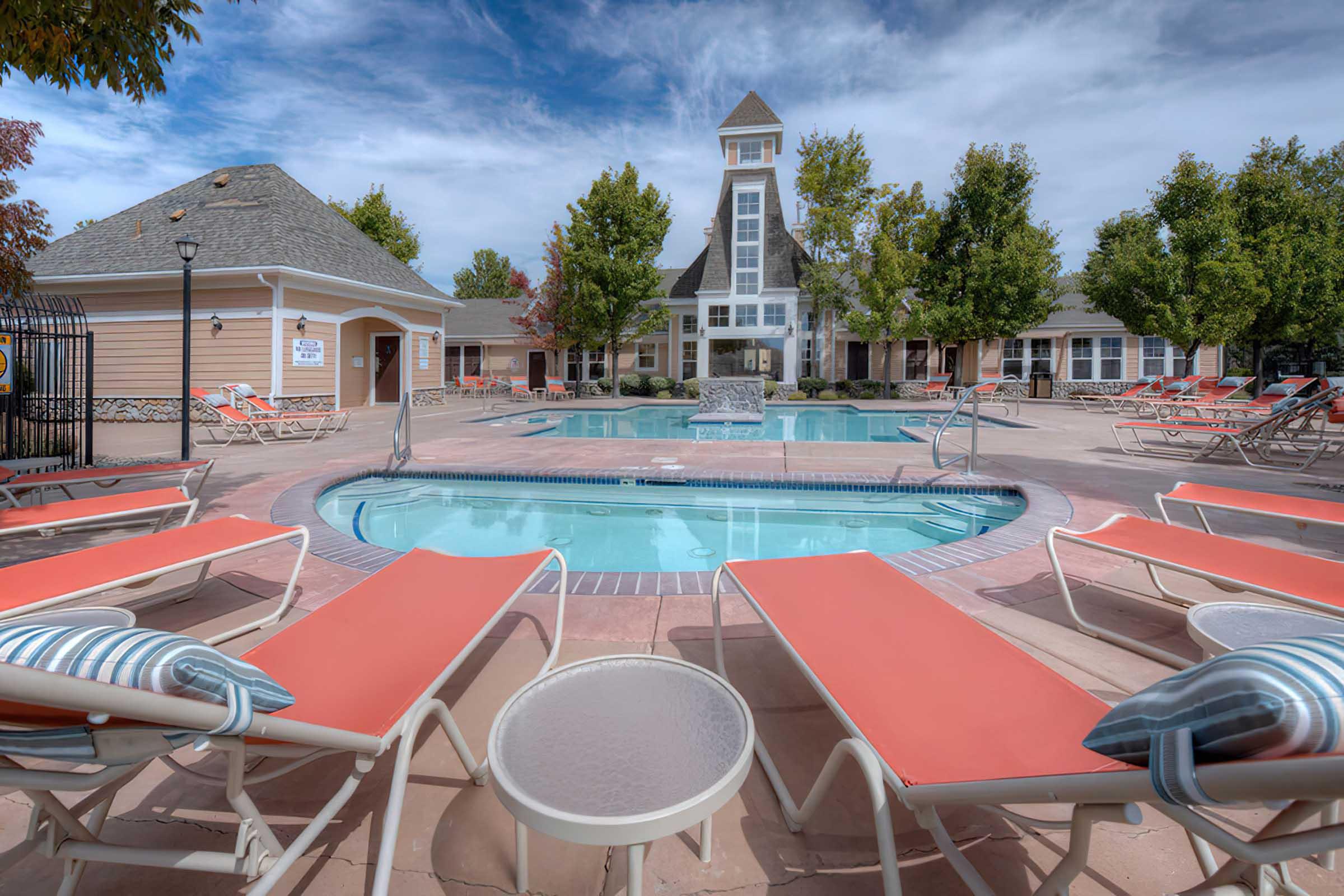 A sunny outdoor pool area surrounded by lounge chairs, with a fountain in the center. The elegant building in the background features a tower and is surrounded by trees. Clear skies above create a welcoming atmosphere for relaxation and leisure.