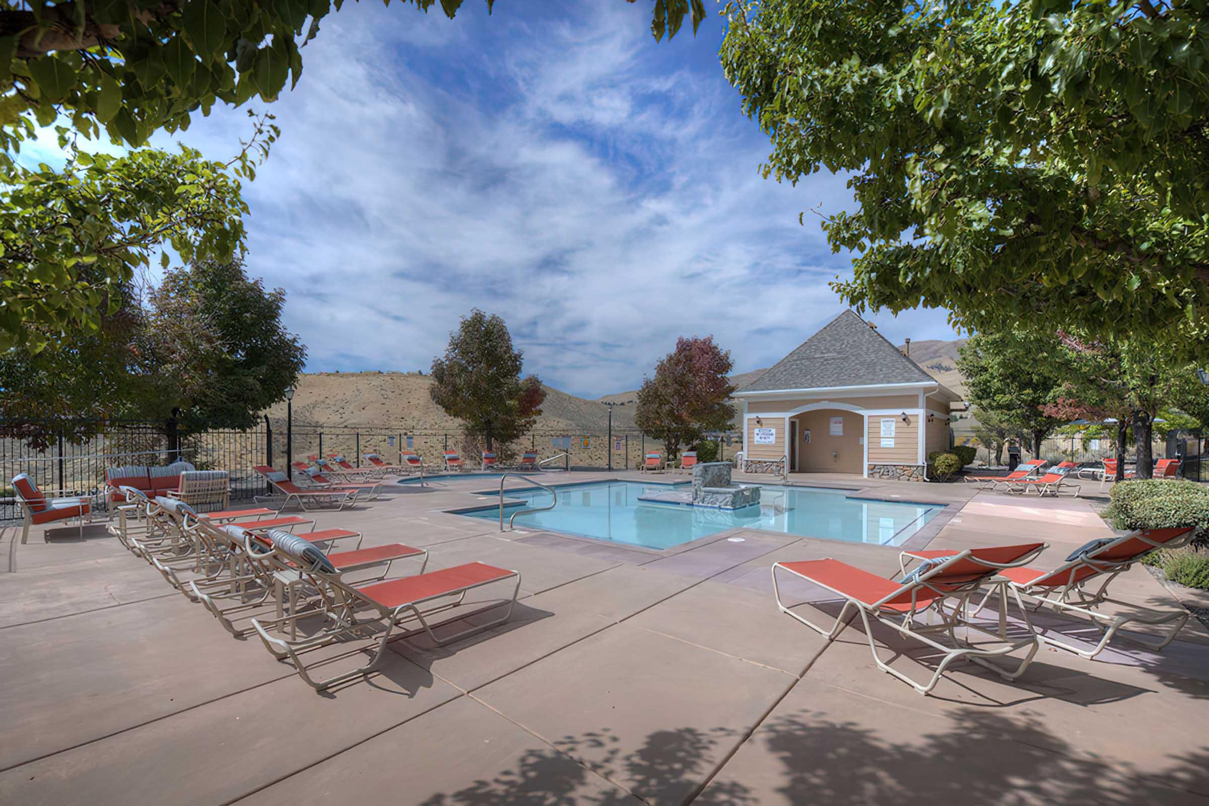 A serene outdoor pool area featuring a clear blue pool surrounded by lounge chairs. Lush trees provide shade, and the sky is partly cloudy. A clubhouse is visible in the background, creating a relaxed atmosphere perfect for leisure and relaxation.