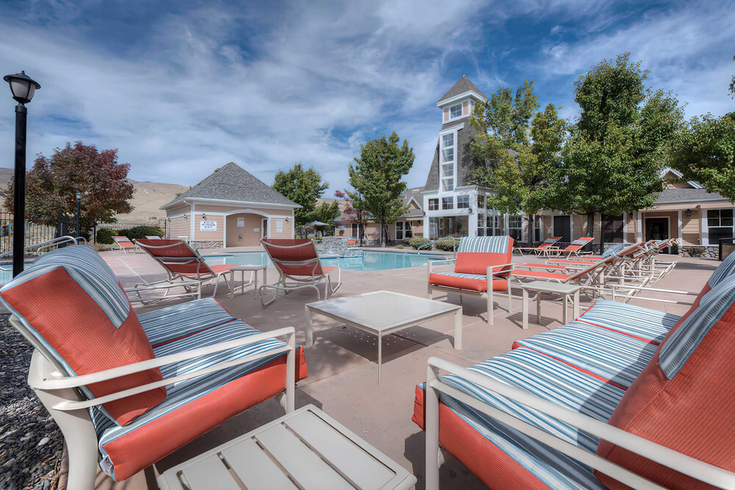 A spacious outdoor pool area surrounded by lounge chairs with orange cushions. In the background, a modern building features large windows and a tall tower. Lush trees and clear blue skies create a serene atmosphere, making it an inviting space for relaxation and leisure.