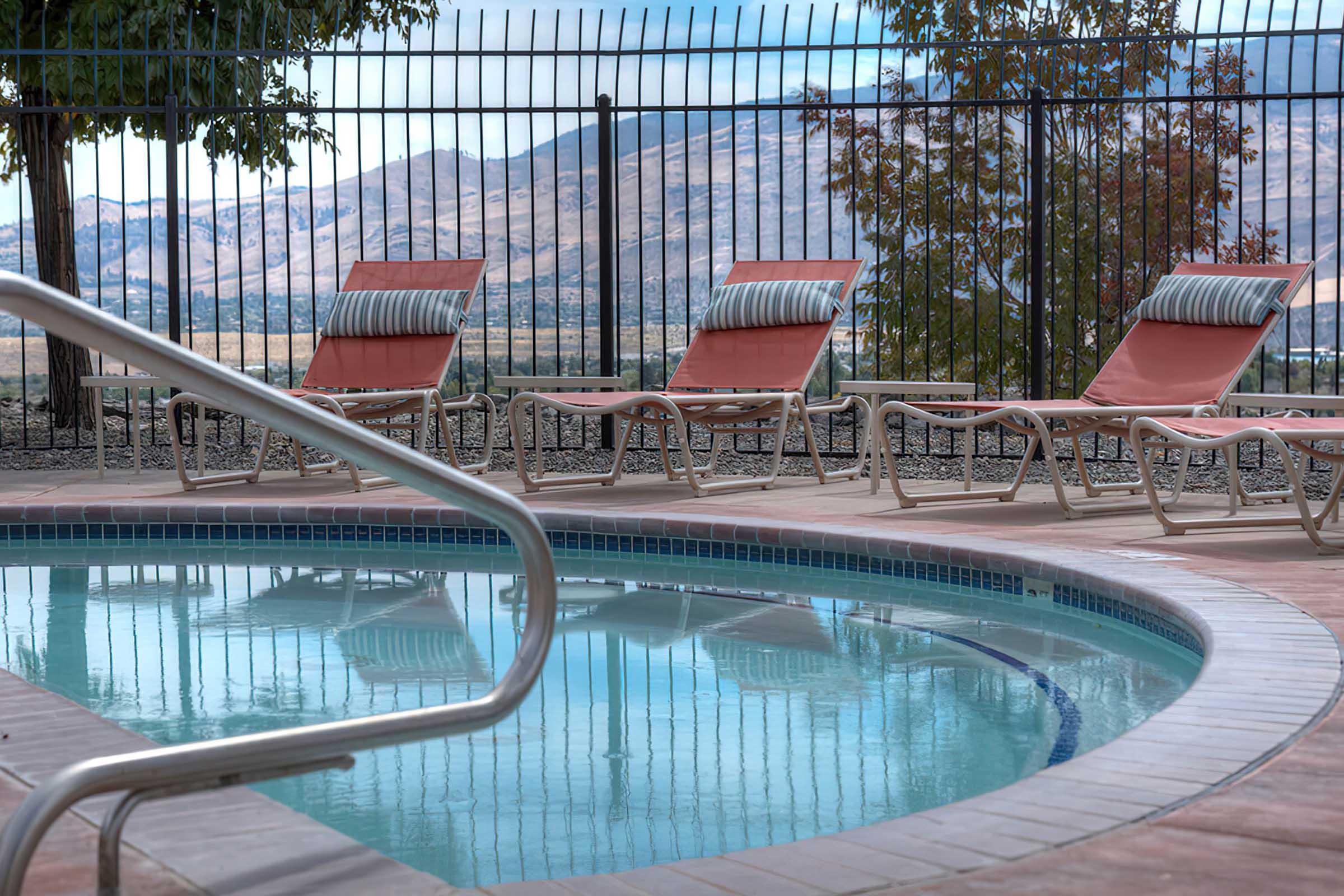 A serene pool area featuring a clear blue swimming pool, surrounded by lounge chairs with red cushions. In the background, a scenic view of mountains and a fence adds to the tranquil atmosphere, creating an inviting space for relaxation.