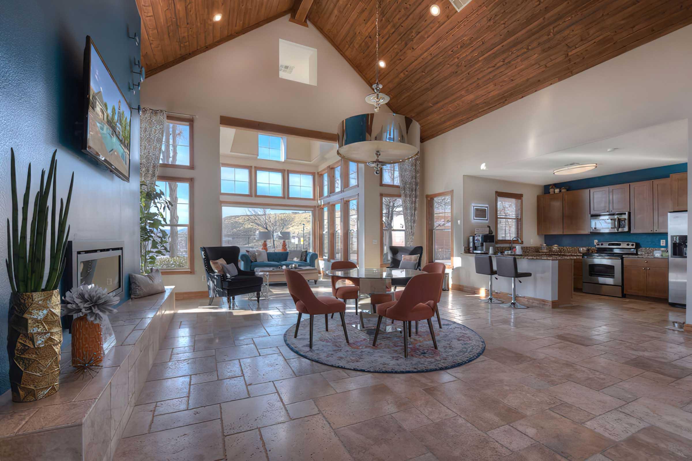 A spacious interior living area with a high wooden ceiling, large windows allowing natural light, and modern furnishings. It features a round rug, a dining table with chairs, a cozy seating area, and a kitchen visible in the background. Potted plants and artwork add a decorative touch to the space.
