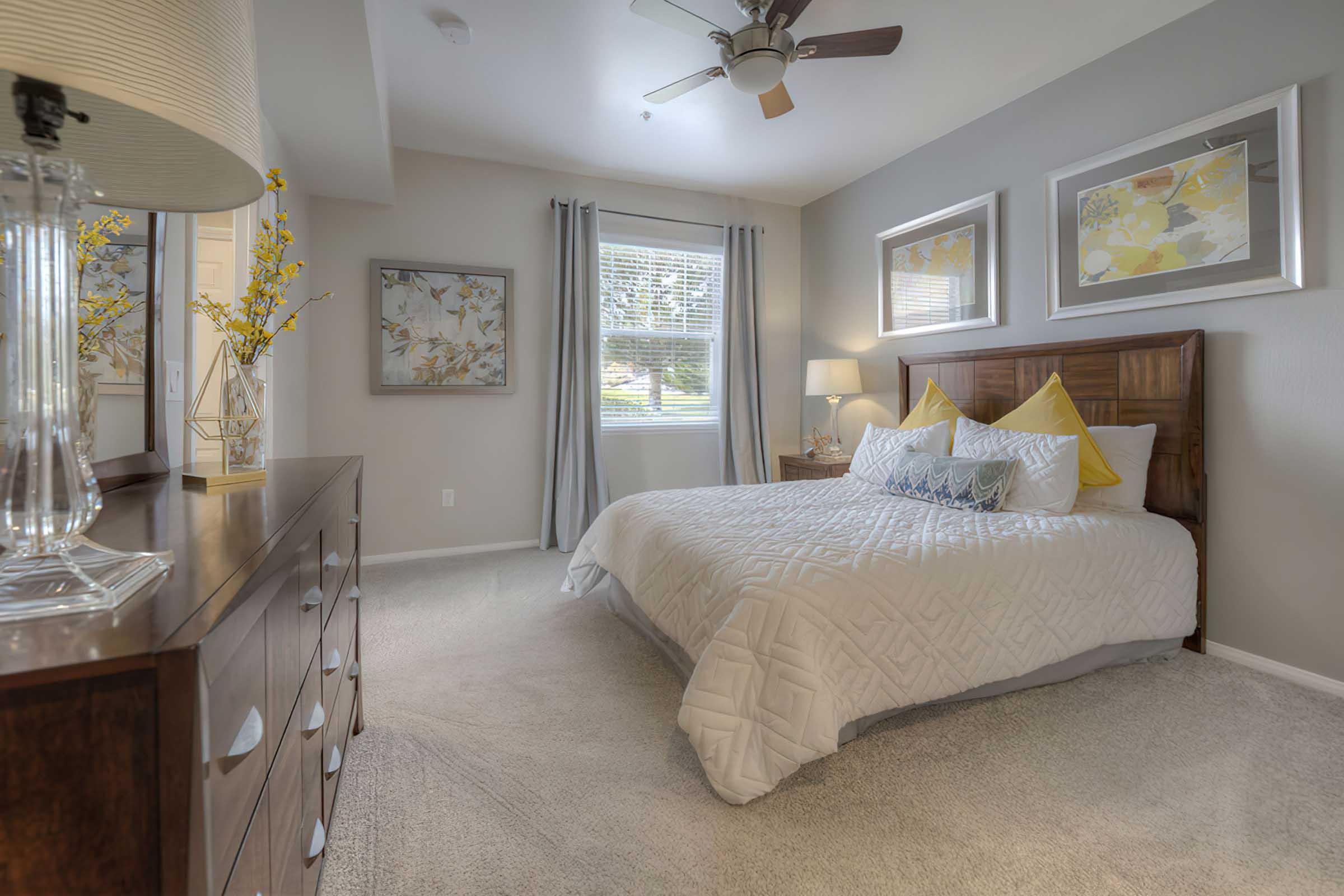 A cozy bedroom featuring a queen-sized bed with a white quilt, yellow accent pillows, and framed artwork on the walls. There is a wooden dresser with drawers, a lamp, and a ceiling fan. Natural light streams through the window adorned with gray curtains, and a vase with yellow flowers adds a touch of color.