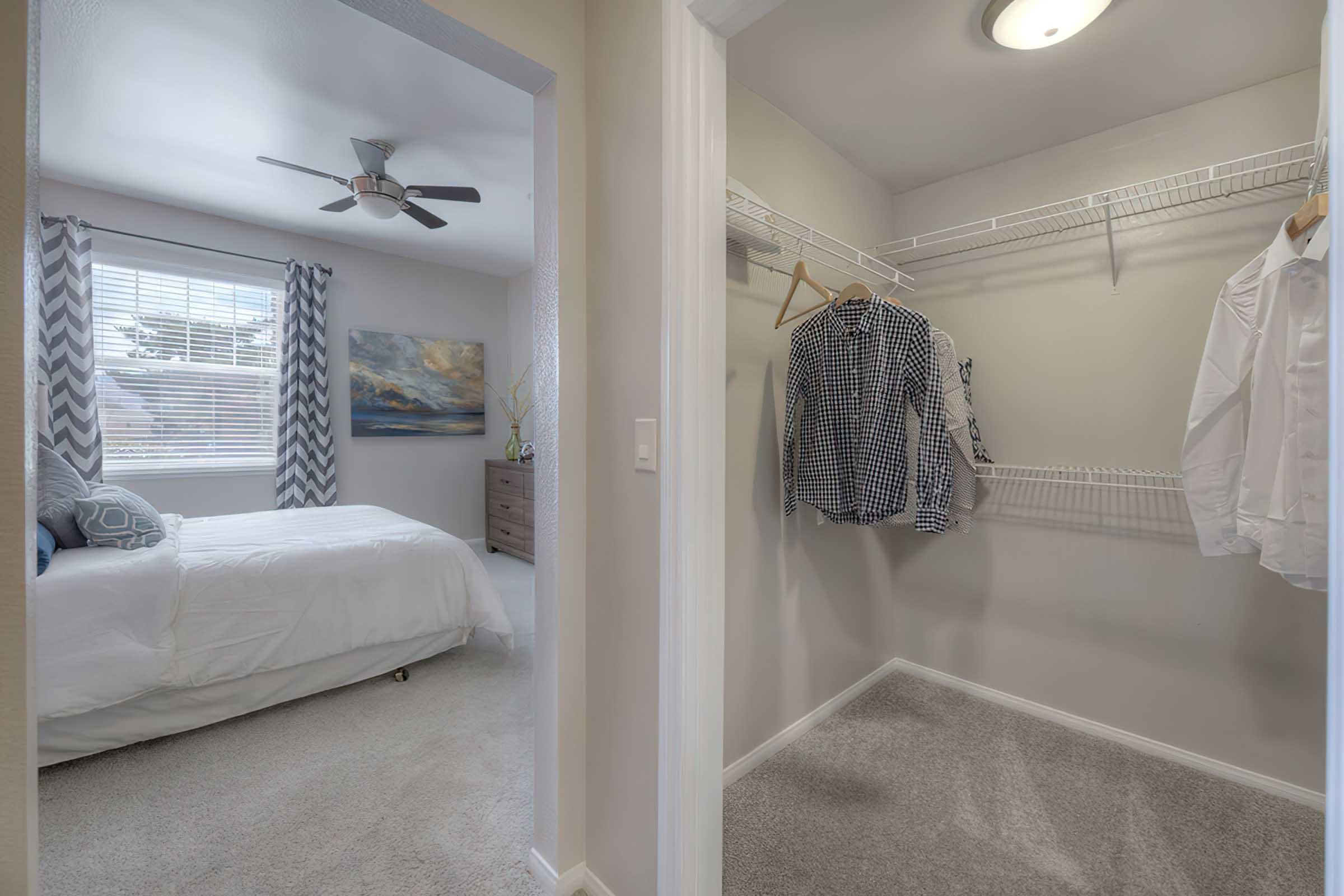 A view of a well-lit room with a bed, decorative pillows, and large windows featuring sheer curtains. In the foreground, an open closet with a white shirt and a patterned shirt hanging on a wire rack. The room has light-colored walls and carpet, creating a clean and spacious atmosphere.