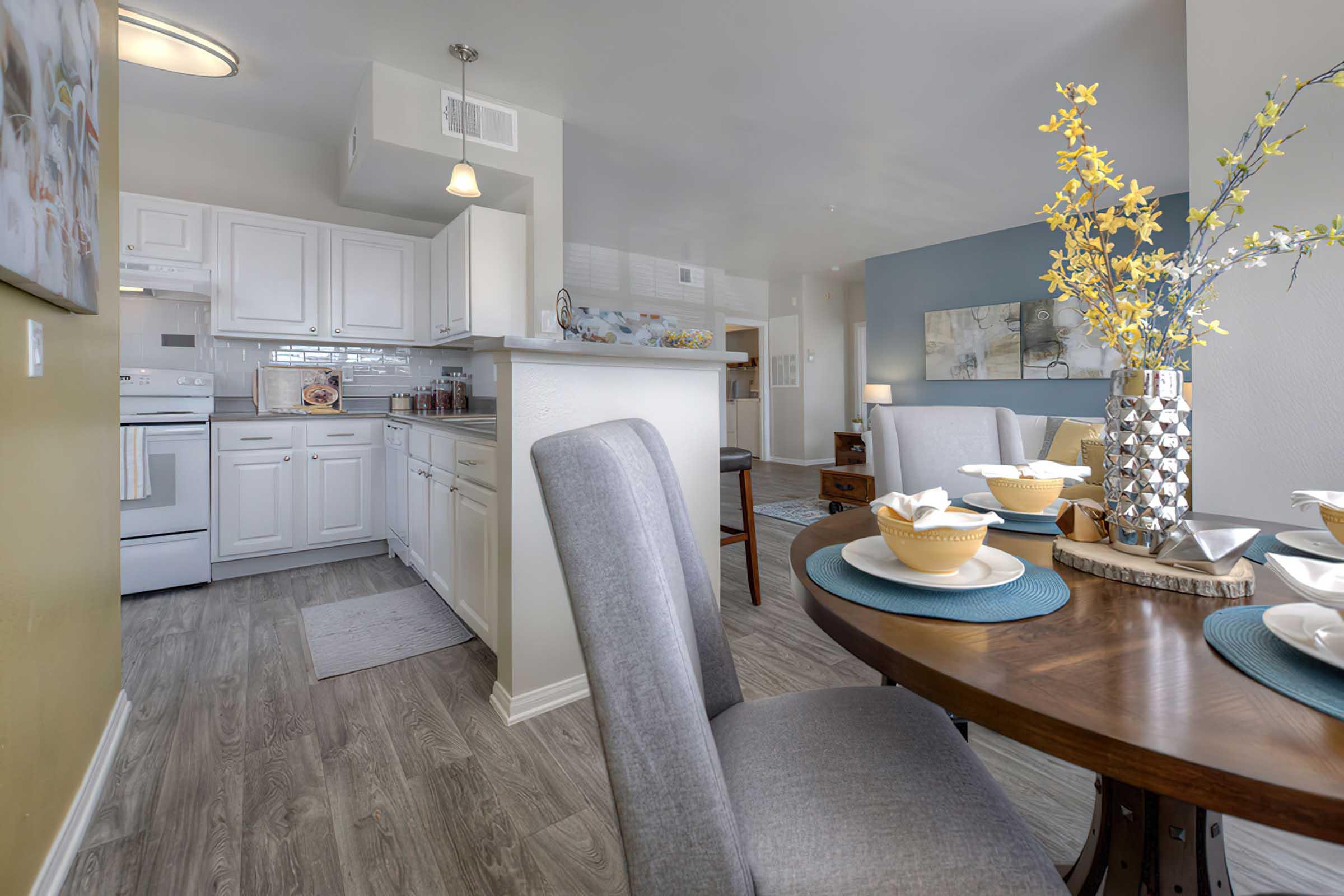 A modern dining area featuring a wooden table set with dishes and decorative items. In the background, a bright kitchen with white cabinetry and stainless appliances is visible. The room has light, neutral colors and is decorated with fresh flowers, creating an inviting atmosphere.