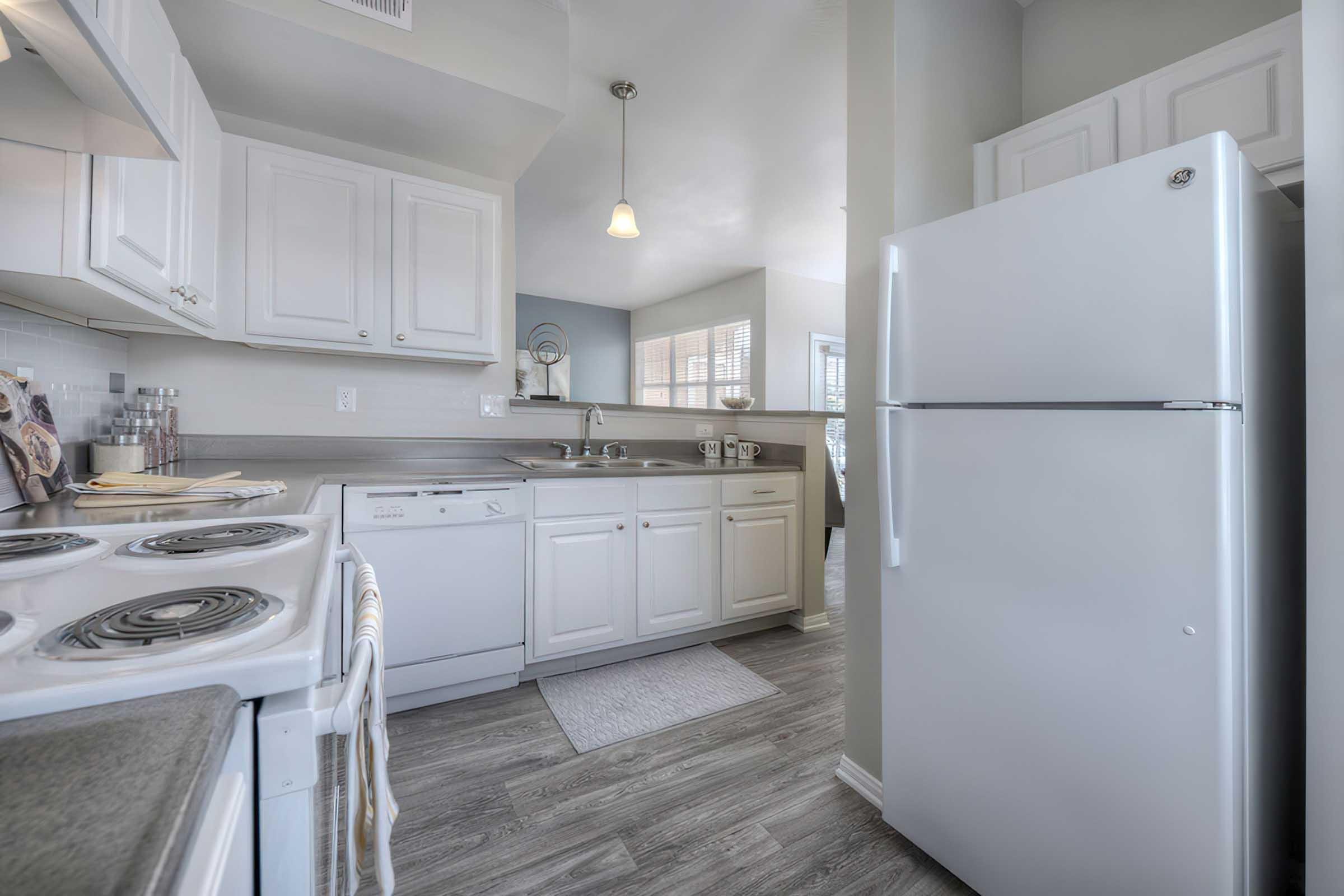 A modern kitchen featuring white cabinets, a stainless steel stove, a dishwasher, and a refrigerator. The room is well-lit, with a window that shows natural light and a small dining area visible in the background. The floor is styled with light wood-like materials, enhancing the spacious and clean aesthetic.