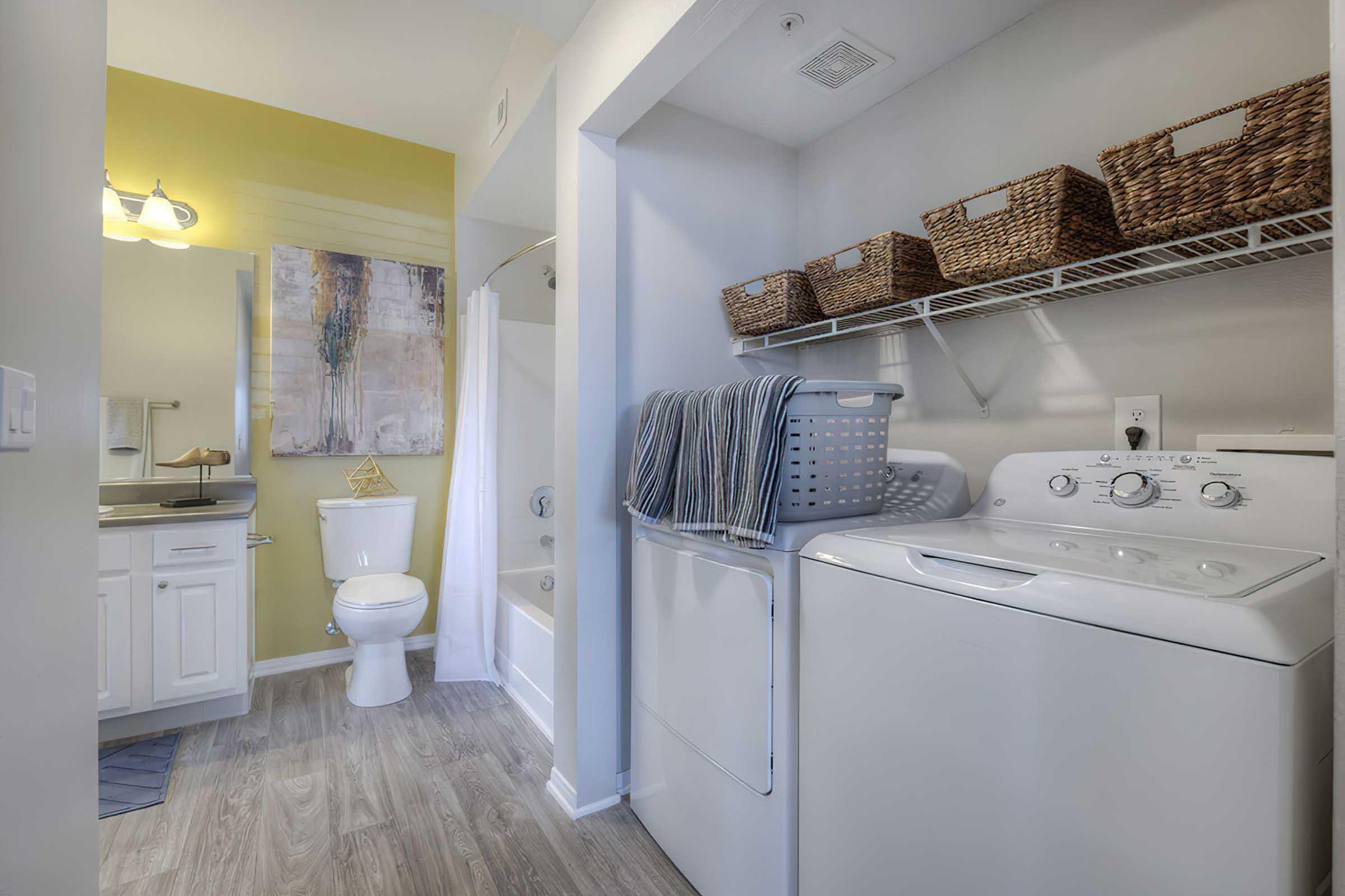 A modern bathroom and laundry area featuring a washer and dryer, a toilet, and a bathtub. The walls are painted a light color with one yellow accent wall. Decorative baskets are placed on a shelf, and there is a mirror and art piece on the wall, creating a functional and stylish space.