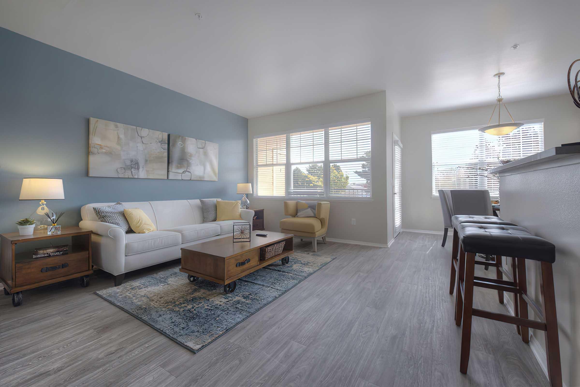 Modern living room featuring a white sofa with yellow and blue accent pillows, a wooden coffee table, and an area rug. Large windows allow natural light, and a cozy armchair is positioned near them. A dining area is visible with barstools and a light fixture above a table, creating an inviting atmosphere.