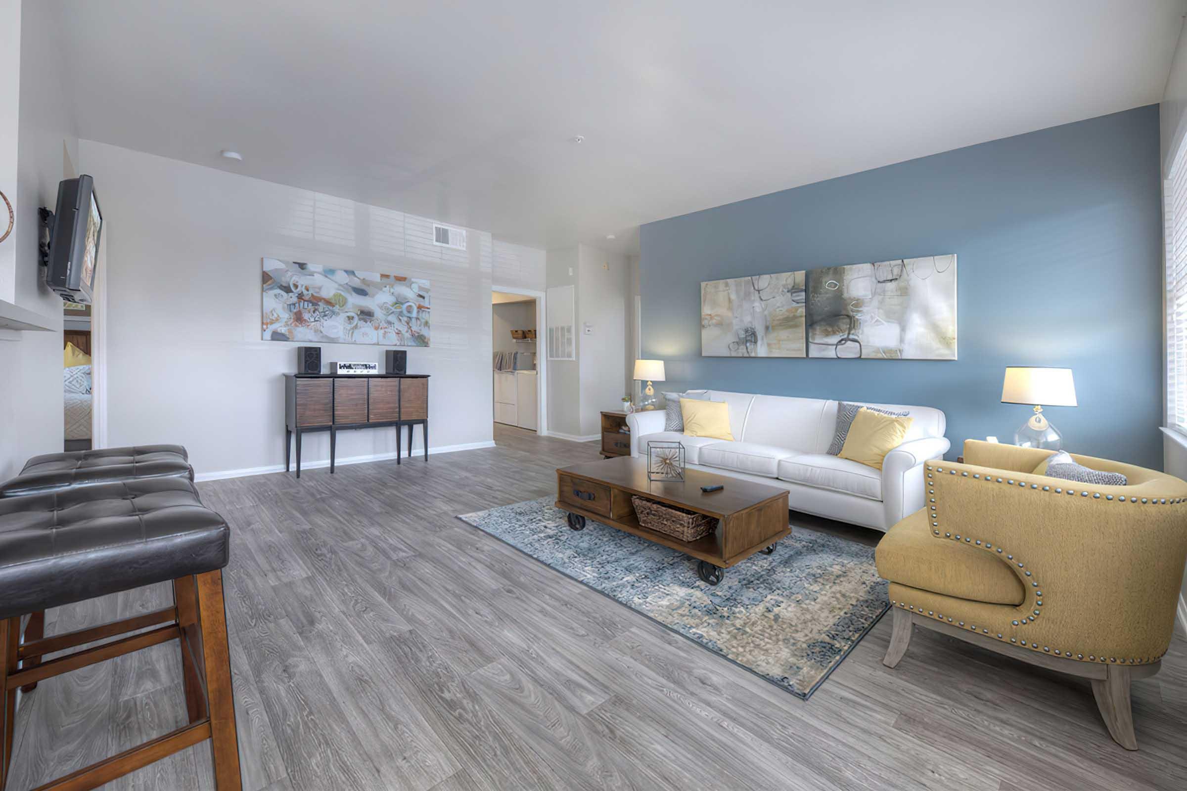 A modern living room featuring a white sofa, a wooden coffee table, and a yellow accent chair. The room has light gray walls and a blue accent wall. There's a flat-screen TV on the wall, a side table, and a decorative rug on the wooden floor. Soft lighting creates a warm and inviting atmosphere.
