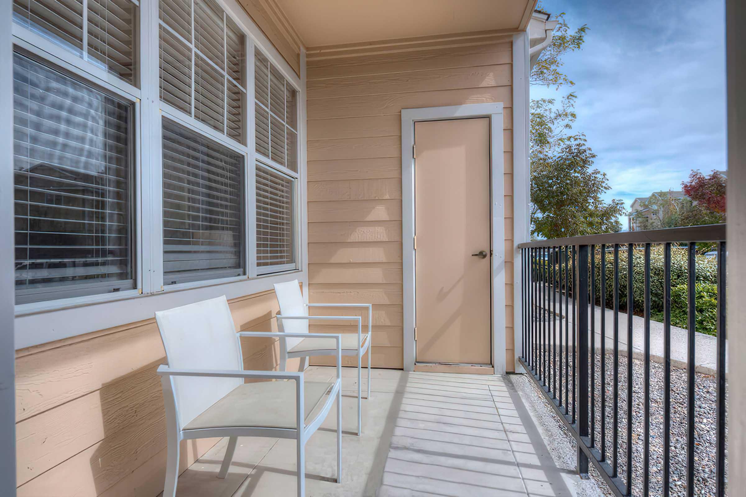 A small outdoor patio area featuring two white chairs and a glass table, situated next to a door. The surrounding walls are painted in a light color, and there are large windows with blinds. The scene is bright and inviting, with greenery visible in the background.