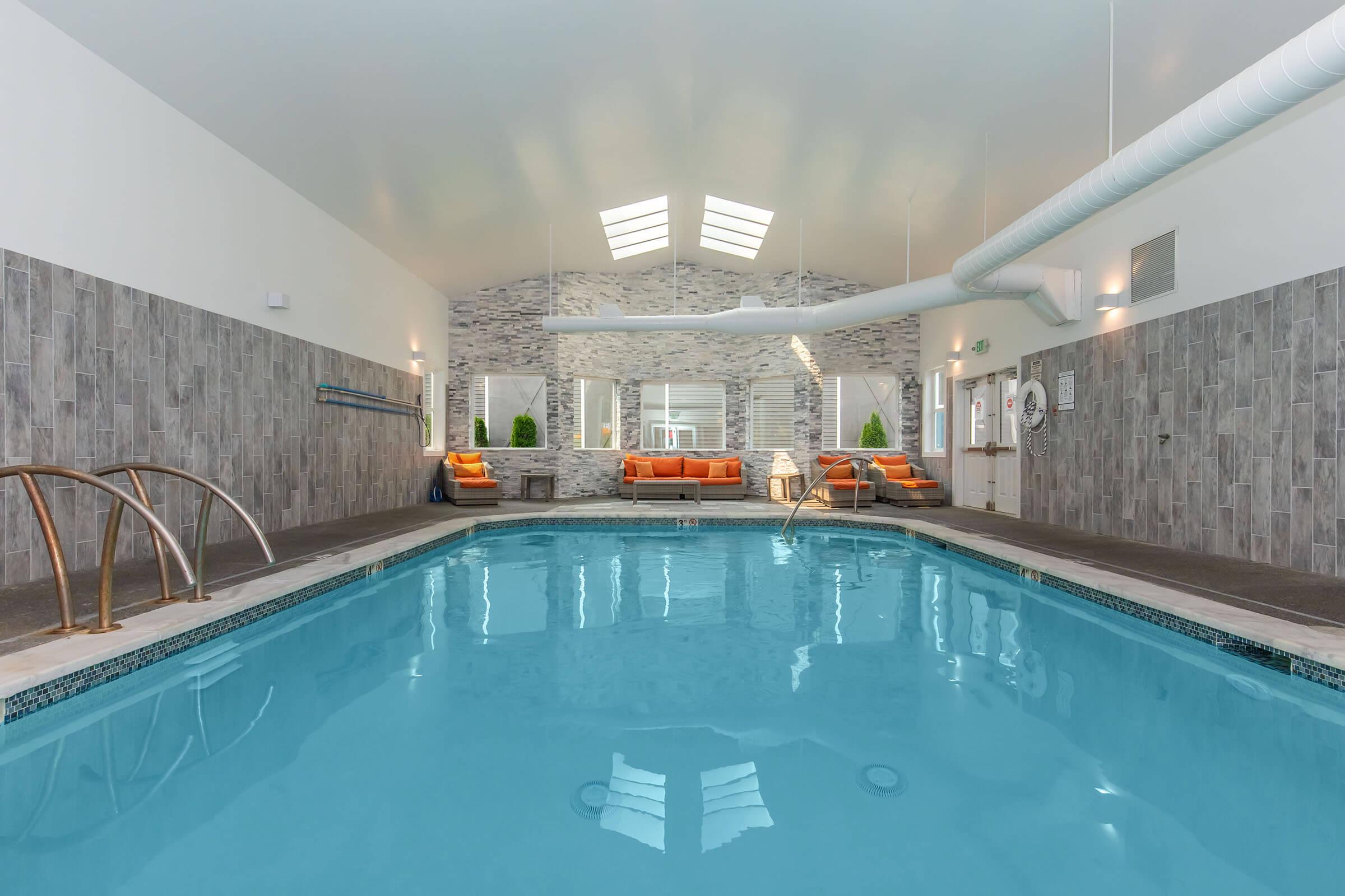 Indoor swimming pool with clear blue water, surrounded by gray stone walls. Natural light enters through skylights and large windows. Comfortable seating with orange cushions is positioned along one wall. The area is bright and inviting, creating a relaxing atmosphere for guests.